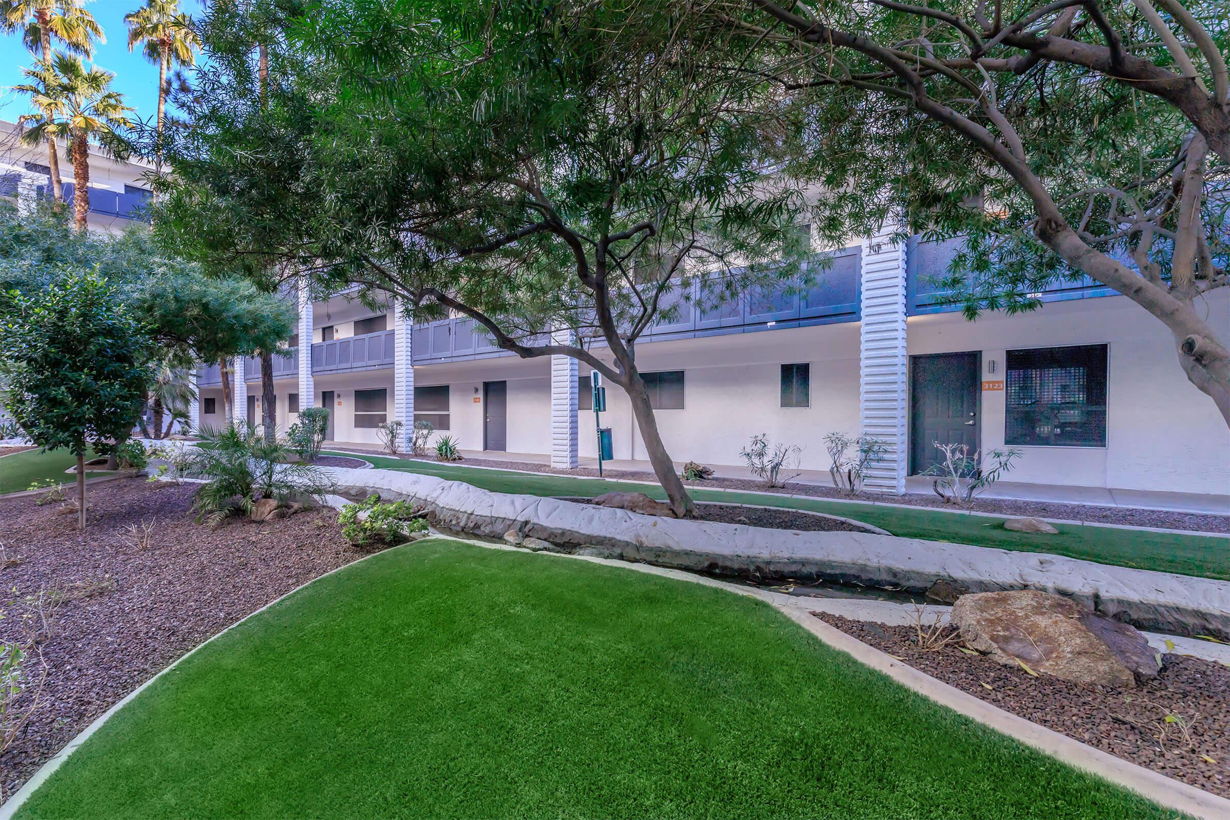 A landscaped courtyard featuring lush green grass, a small stream, and trees, bordered by a white building with balconies.