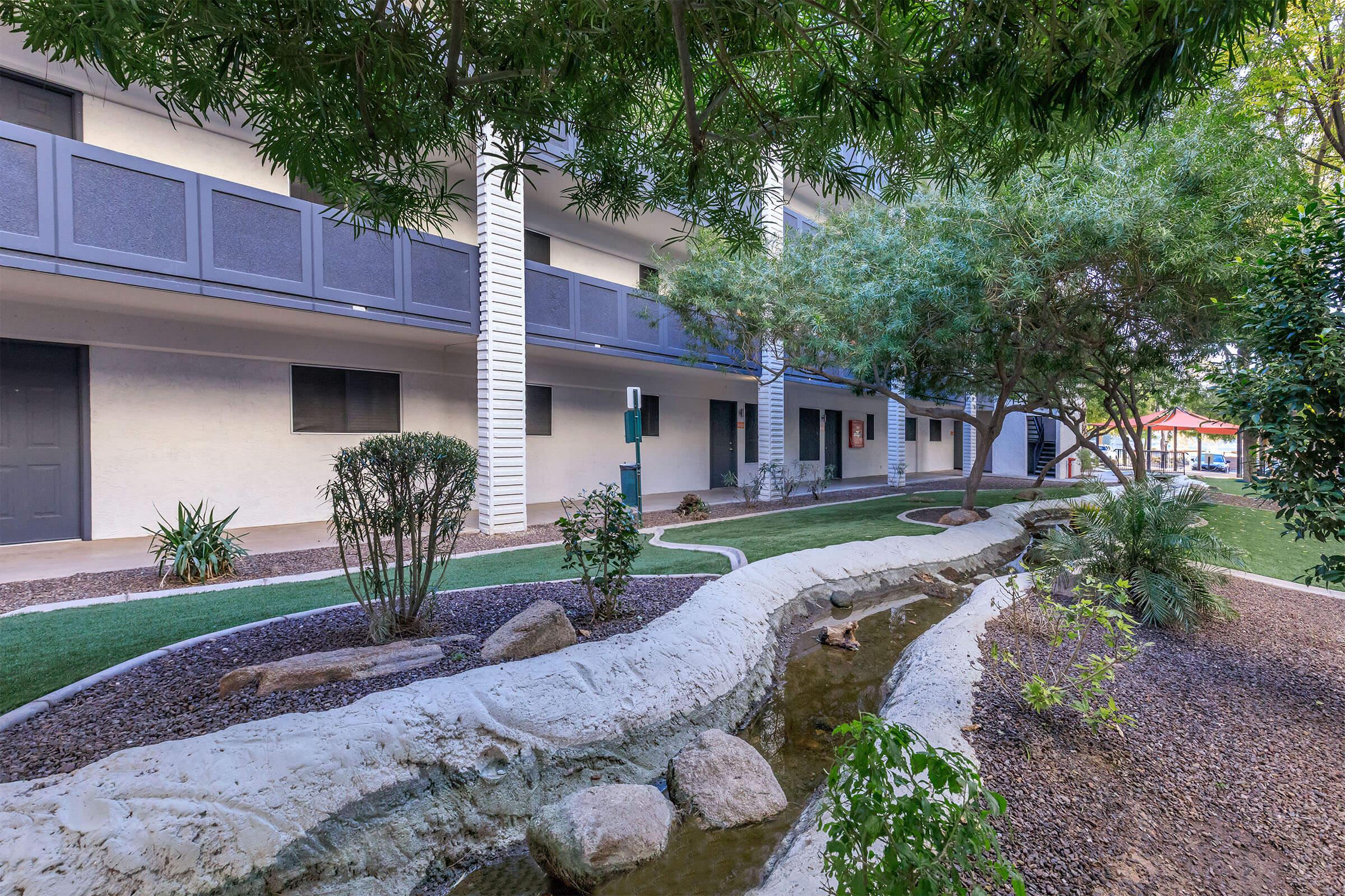 A landscaped courtyard featuring a small stream or water feature, surrounded by greenery, shrubs, and decorative rocks. There are multiple buildings with balconies in the background, and a well-maintained grassy area.