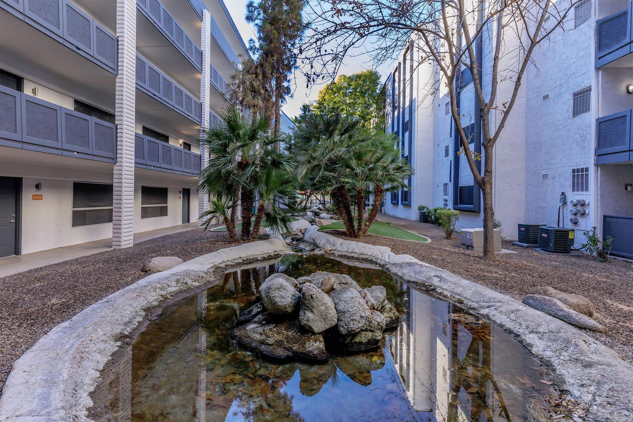 A peaceful courtyard featuring a pond with rocks, surrounded by palm trees, with two multi-story buildings on either side. The scene is bathed in natural light, creating a serene outdoor space.