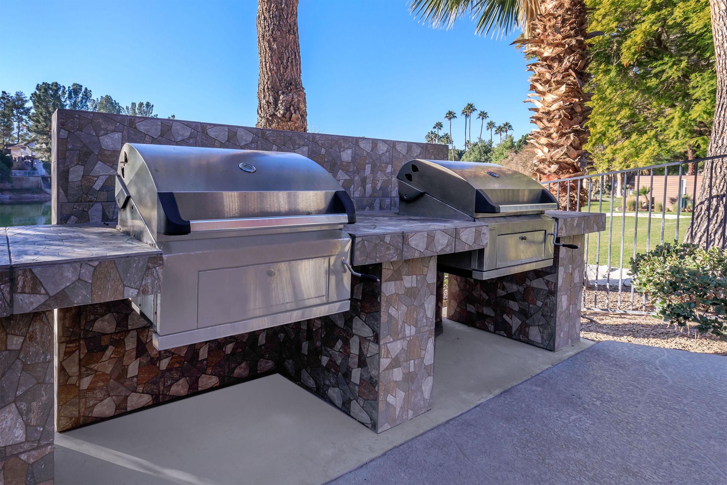 Two stainless steel grills set on a stone mosaic countertop with a palm tree in the background, located in an outdoor area near a body of water.