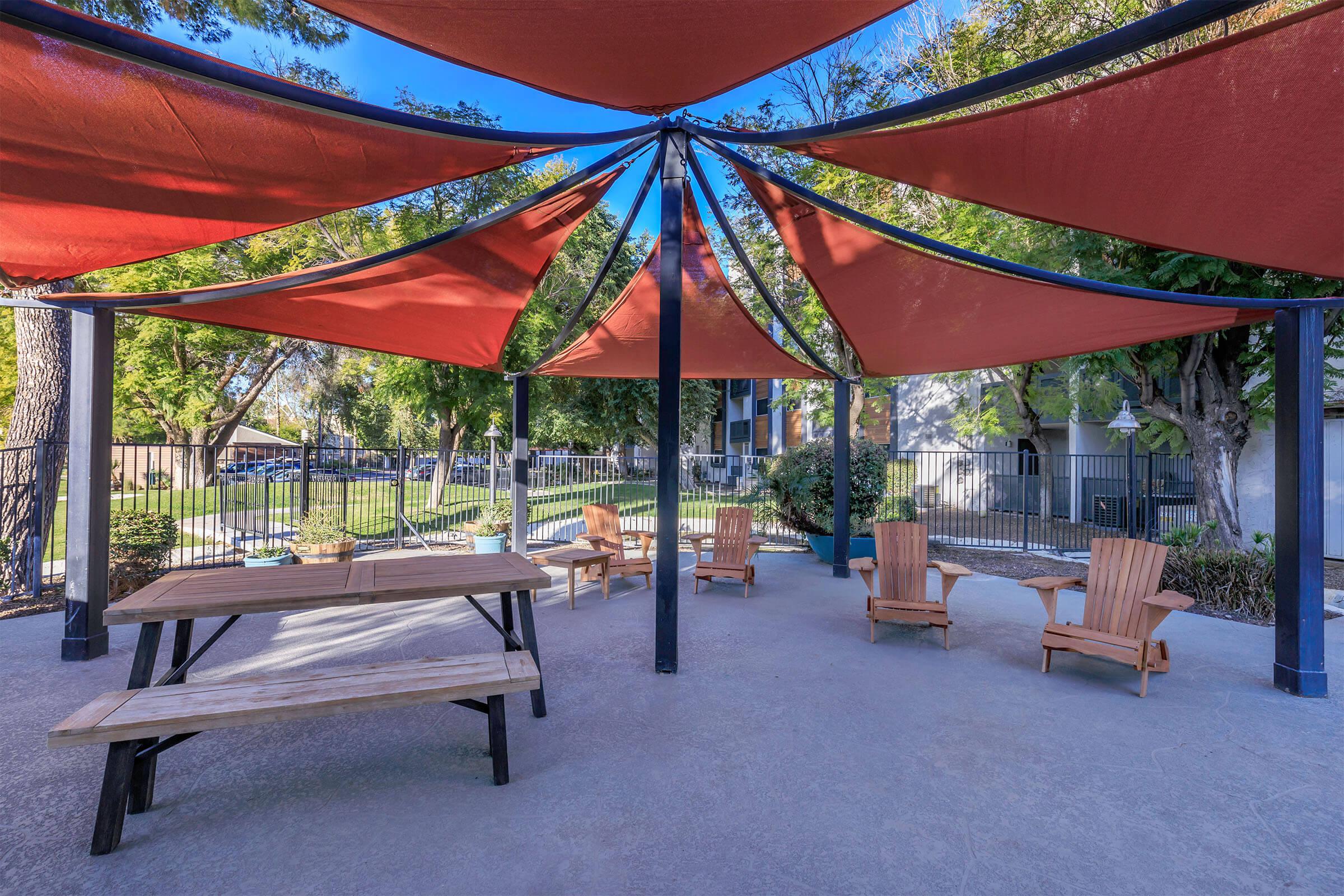 A shaded outdoor seating area with a wooden table and Adirondack chairs, surrounded by greenery and a fence. The structure features a large, colorful fabric canopy supported by black poles.