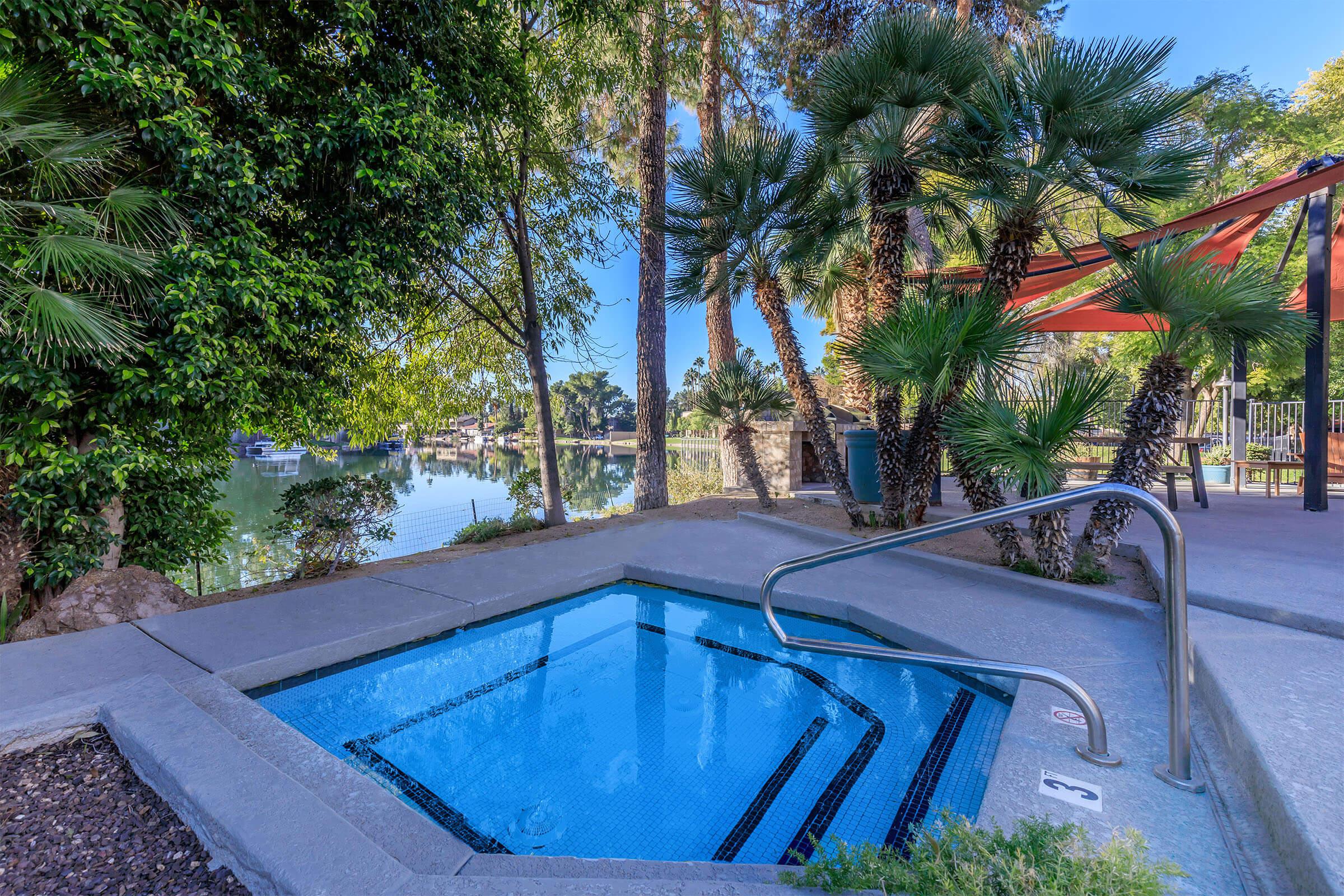 A tranquil outdoor hot tub surrounded by lush greenery and tall palm trees, with a view of a calm water body in the background. Bright sunlight illuminates the scene, creating a relaxing atmosphere.