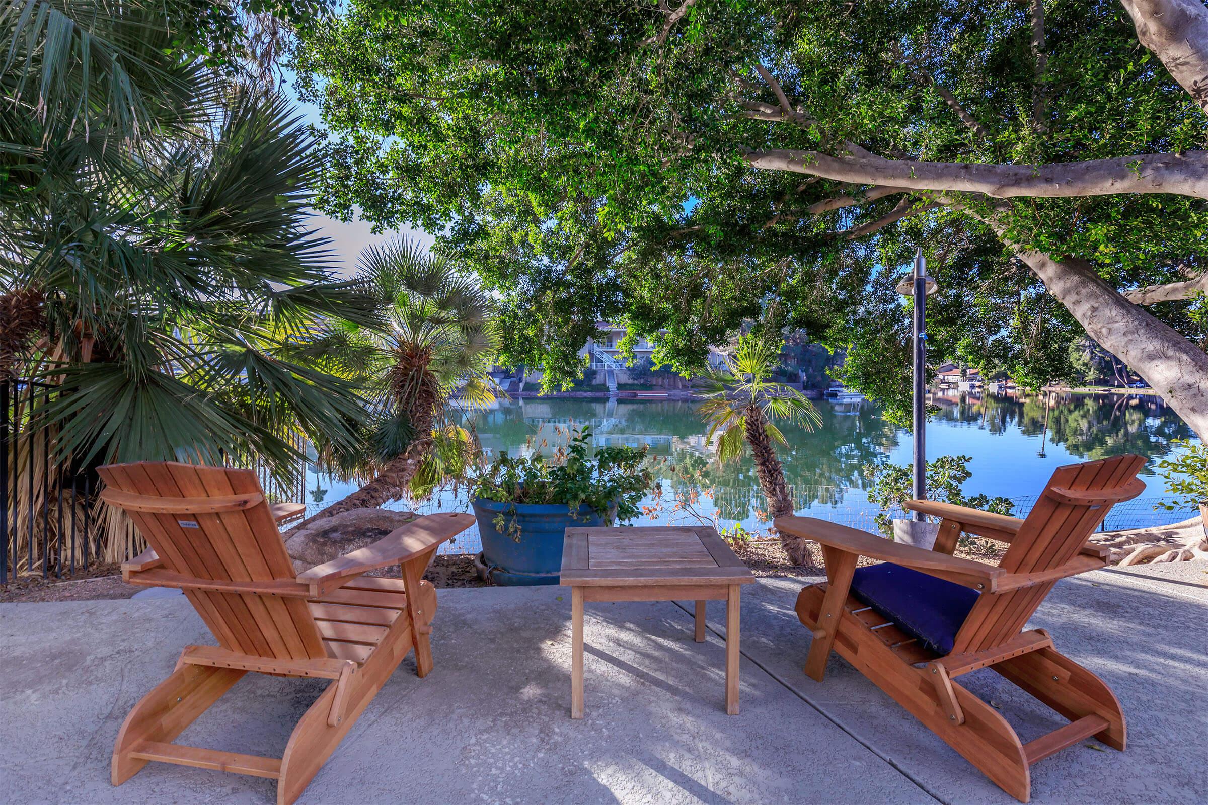 A serene outdoor scene featuring two wooden lounge chairs and a small table overlooking a calm body of water surrounded by lush greenery and palm trees.