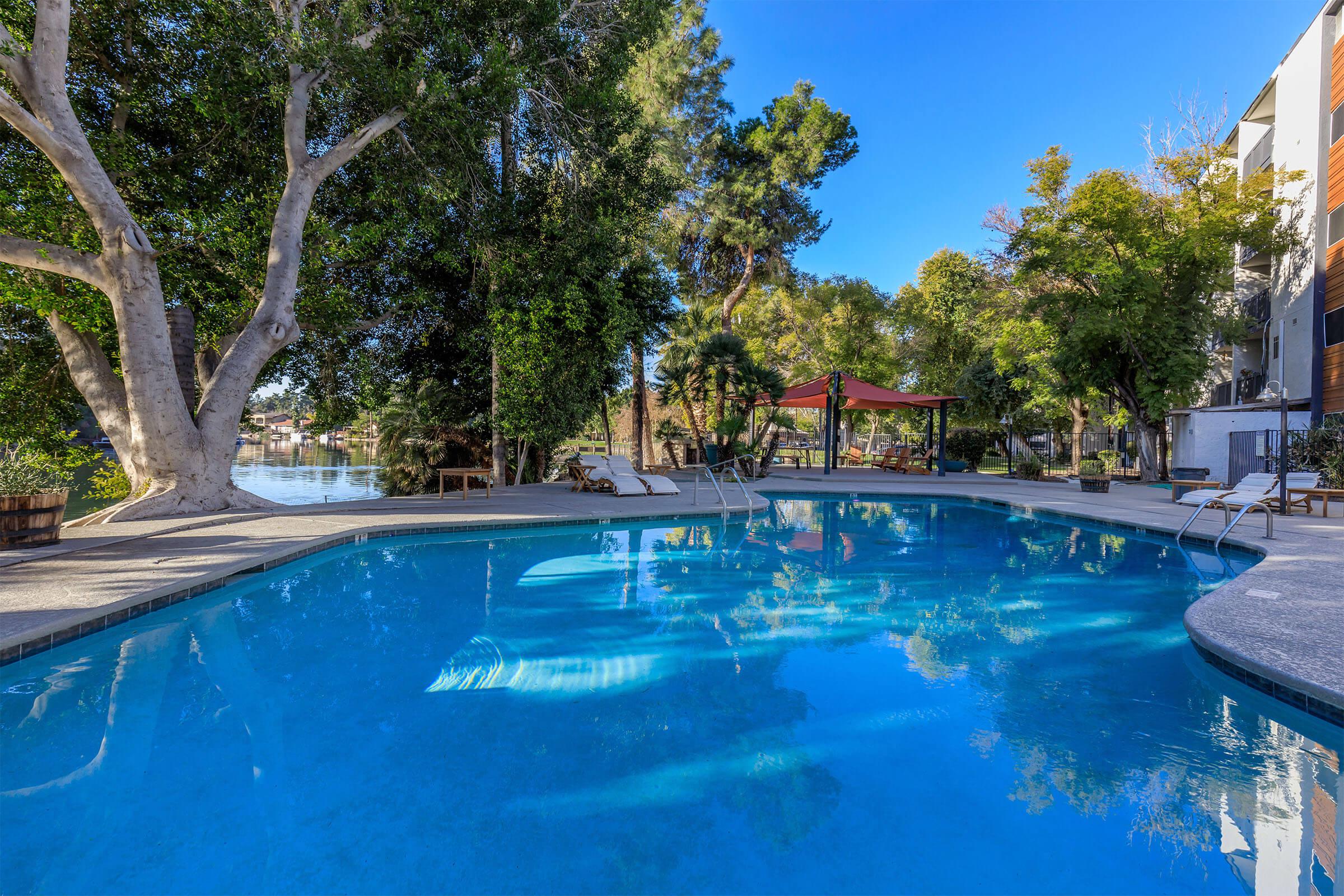 A clear blue swimming pool surrounded by lush greenery, with sun loungers nearby and a shaded area under a gazebo. The scene is bright and inviting, reflecting a serene outdoor environment.