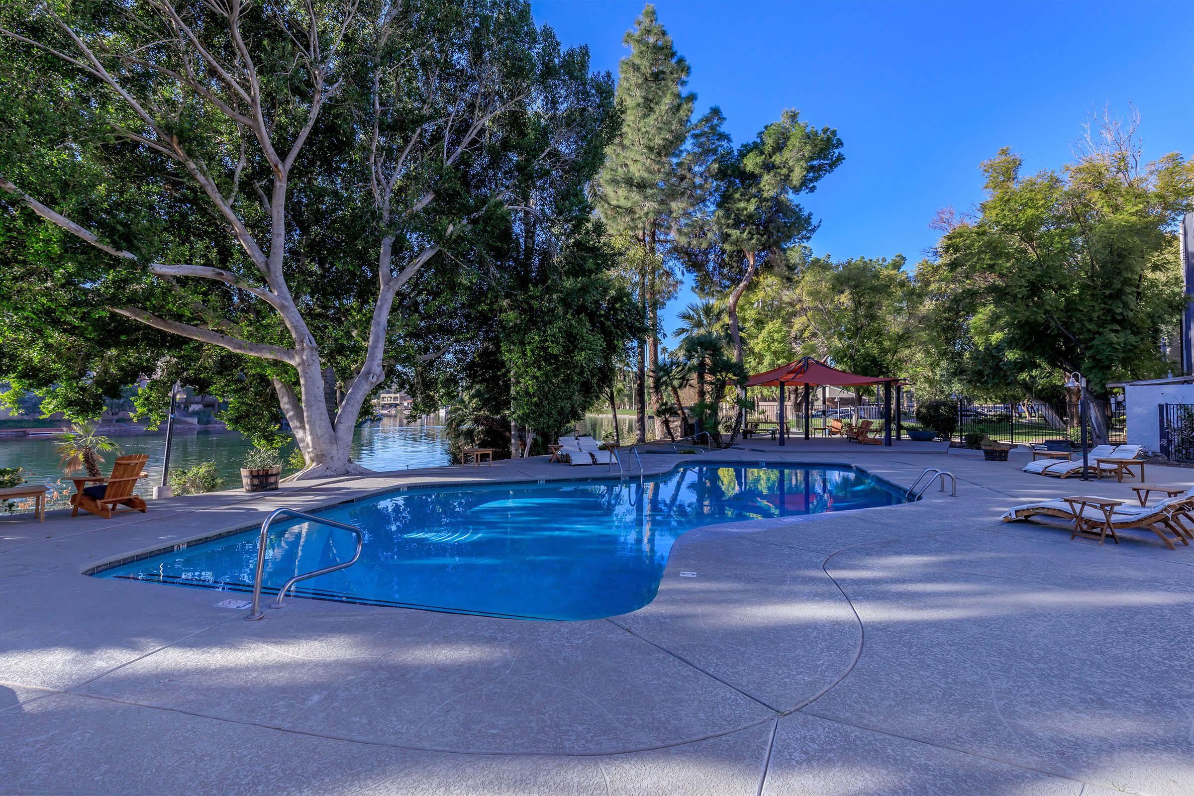 A serene outdoor pool area surrounded by trees, with lounge chairs and a gazebo. The water is clear and inviting, reflecting the blue sky, and the scene offers a peaceful and relaxing atmosphere.