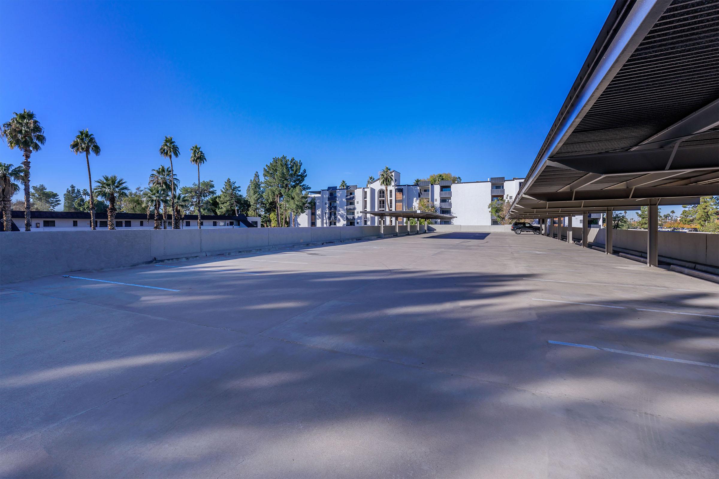 A nearly empty parking lot with palm trees in the background and residential buildings visible. The sky is clear and blue, and the lot features a few shaded parking spaces under a canopy.