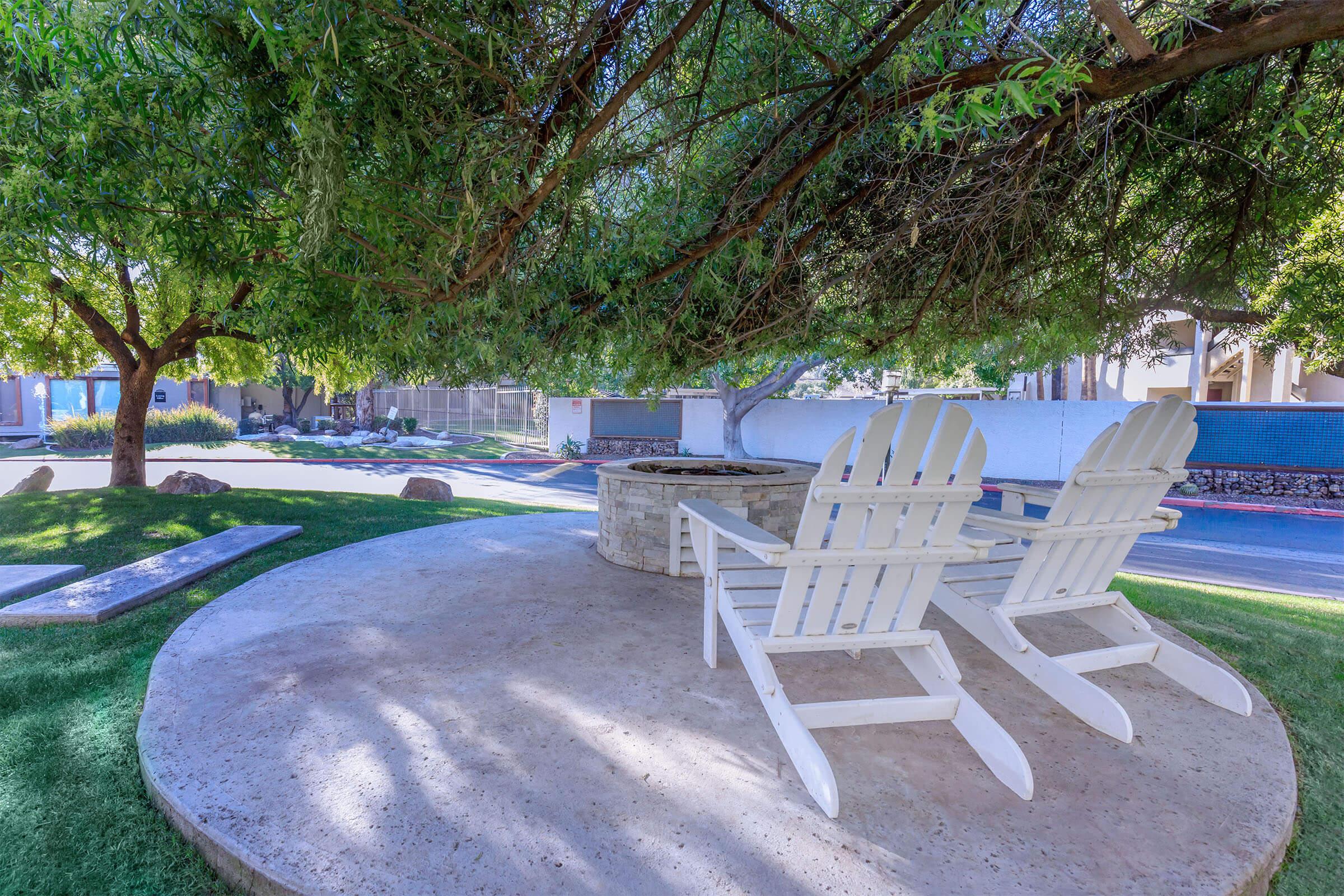 A shaded outdoor seating area featuring two white Adirondack chairs positioned next to a stone fire pit, surrounded by grass and trees.