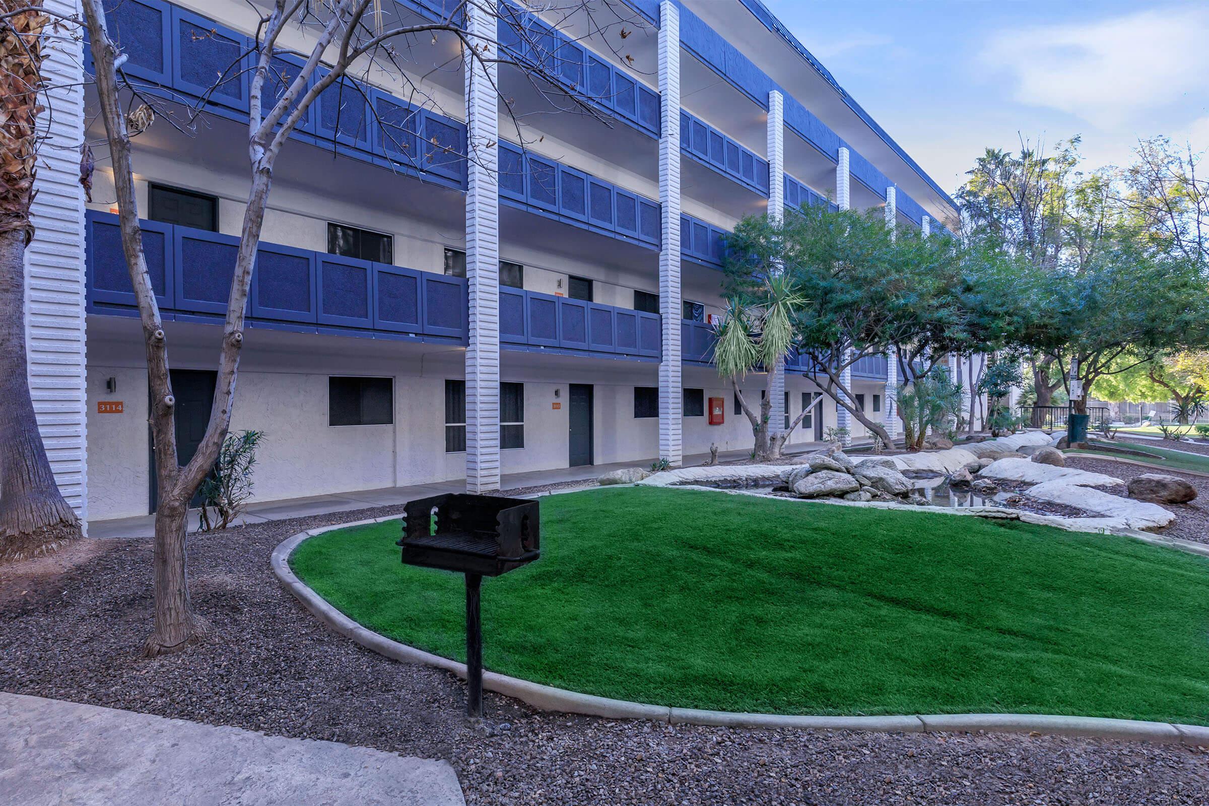 A view of a modern apartment community with multiple balconies, surrounded by green grass and decorative rocks. There are trees and shrubs planted in the area, and a barbecue grill is positioned on a small patio in the foreground.