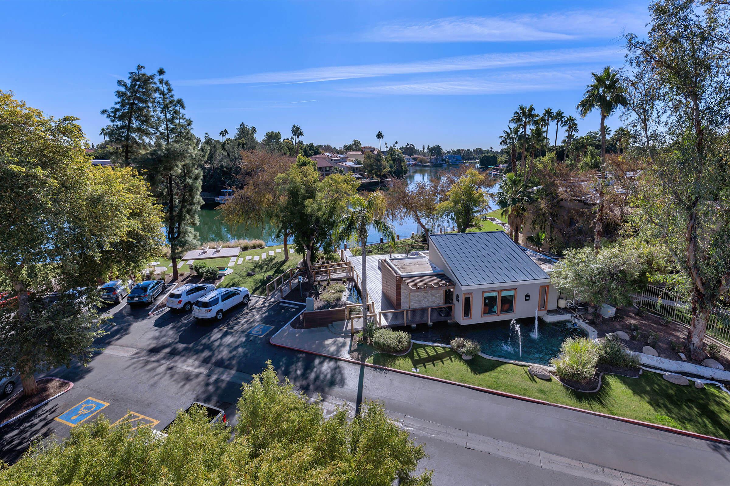 Aerial view of a lakeside building surrounded by palm trees, with a parking area in front. The scene features clear blue skies and a tranquil water setting, creating a serene atmosphere.