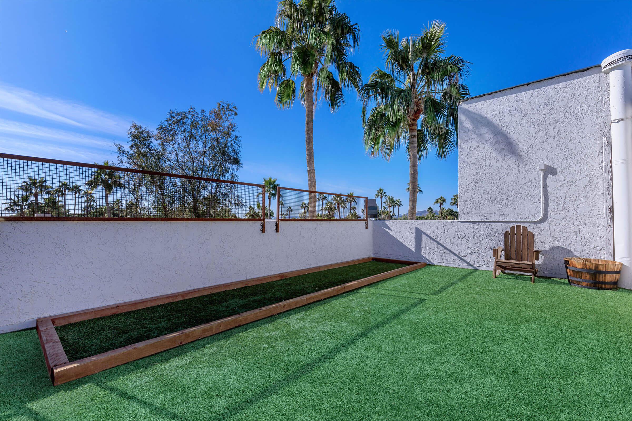 A rooftop area with artificial grass, featuring palm trees in the background, a wooden lounge chair, and a long narrow planting bed along the side.