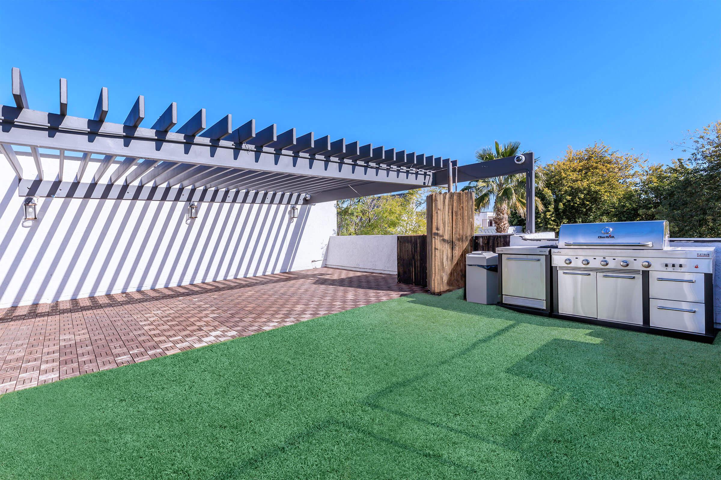 An outdoor patio with artificial grass, featuring a modern stainless steel barbecue grill and a cooking station under a pergola. Bright blue sky and greenery in the background.