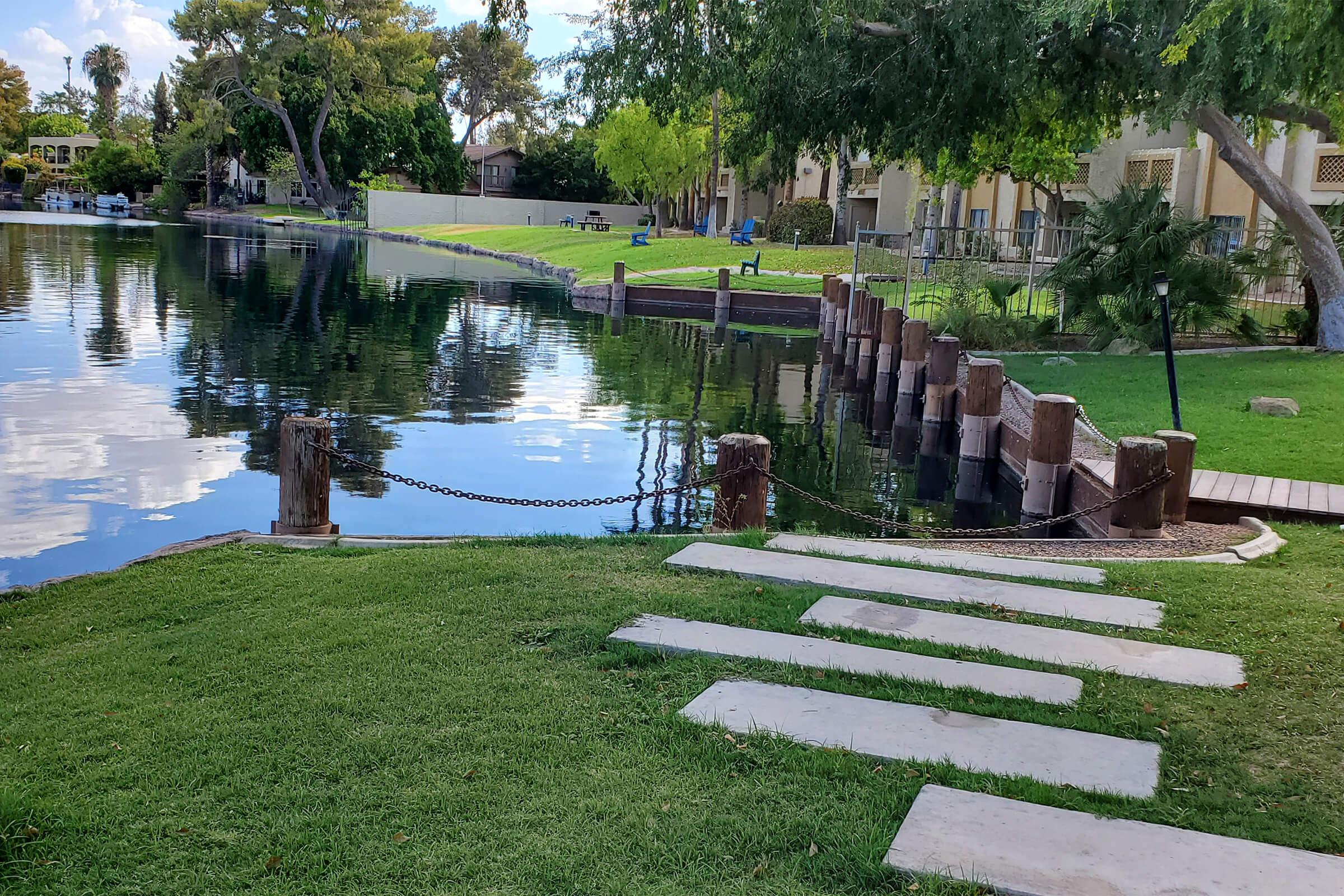 A serene lakeside scene with a grassy area leading to a calm body of water. Wooden posts with a chain separate the lawn from the water. Smooth stone steps descend to the water's edge, surrounded by lush green grass and trees, with a glimpse of a residential area in the background.