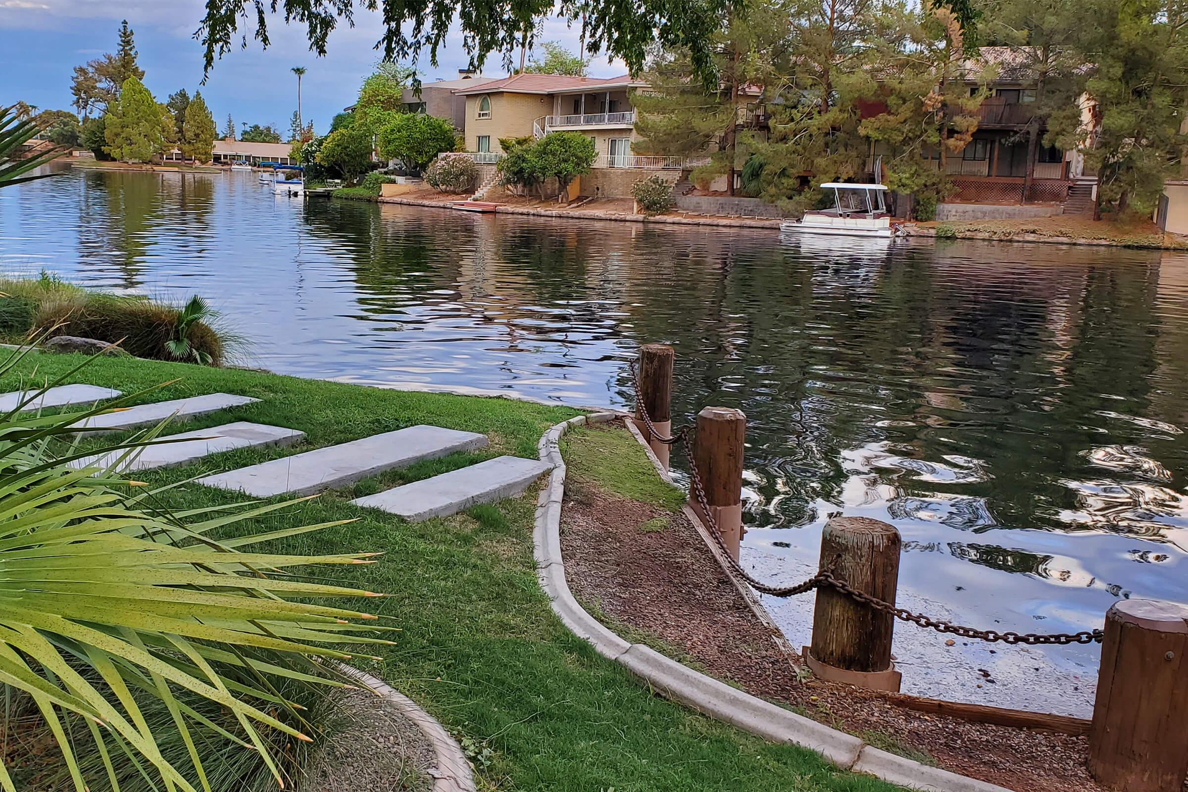 A serene lakeside view featuring grassy banks, wooden posts along the water, and a small boat moored nearby. Trees and house reflections create a tranquil atmosphere, with a pathway leading to the water's edge under a partly cloudy sky.