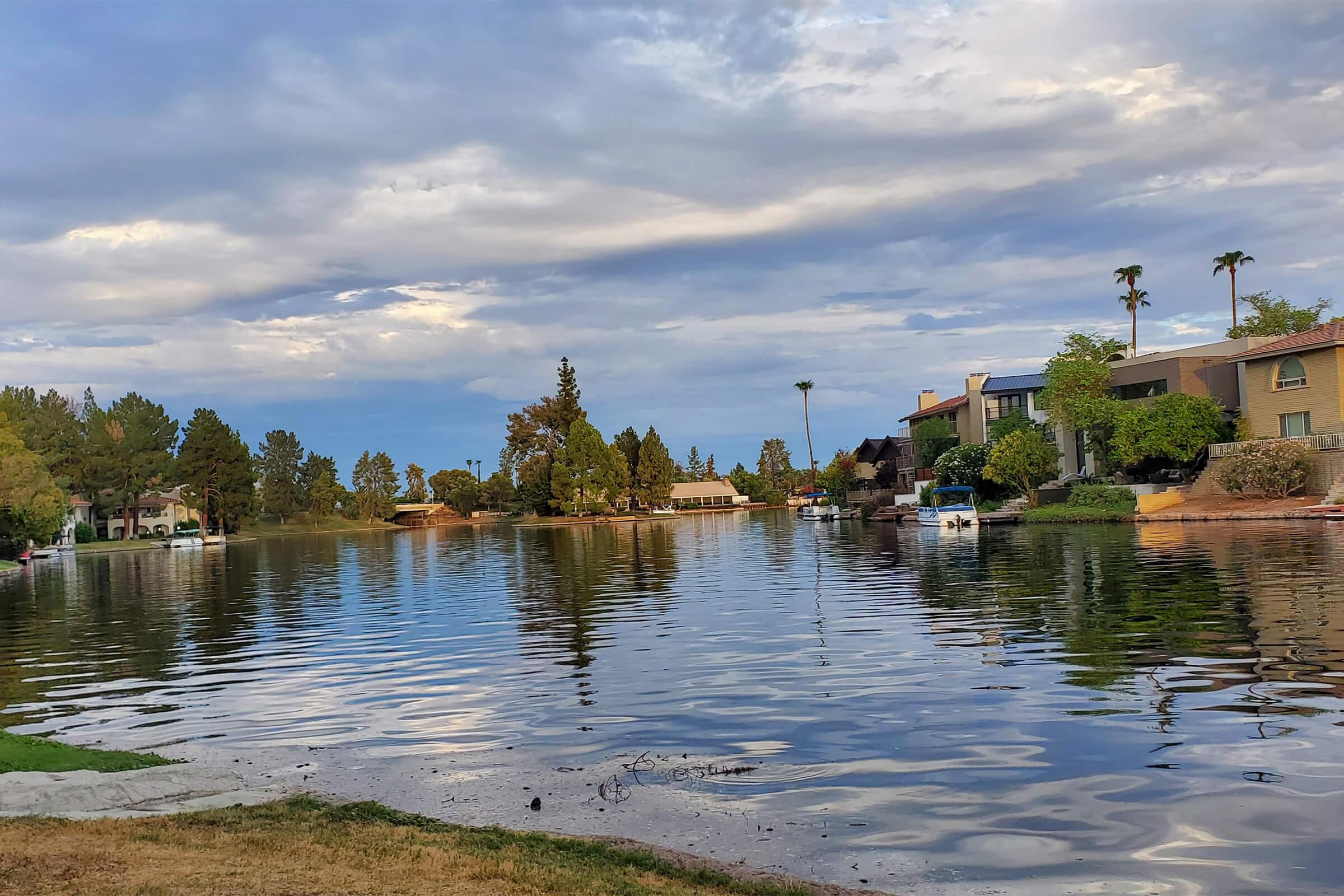 A serene lake scene reflects cloudy skies, surrounded by green trees and residential buildings. The water is calm, with a sandy shore in the foreground. A few boats can be seen on the lake, adding to the tranquil atmosphere of the landscape.