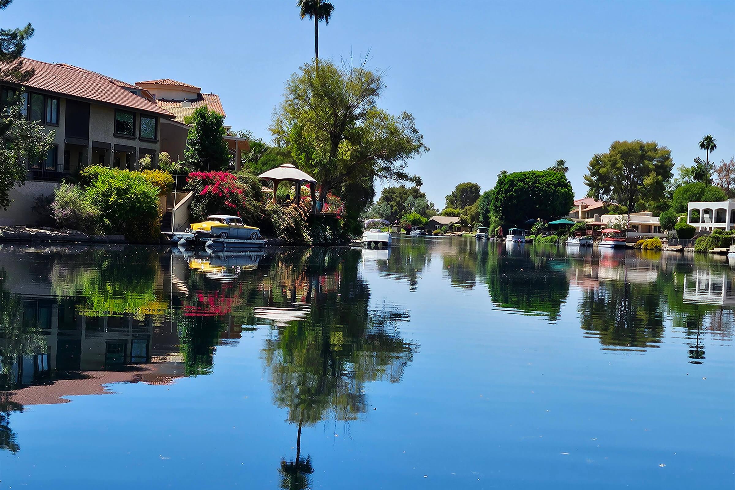A serene canal lined with trees and colorful flowers, reflecting the clear blue sky. Houses with balconies and boats are visible, creating a peaceful waterfront scene under bright sunlight. Palm trees are dotted along the shoreline, enhancing the tranquil atmosphere.
