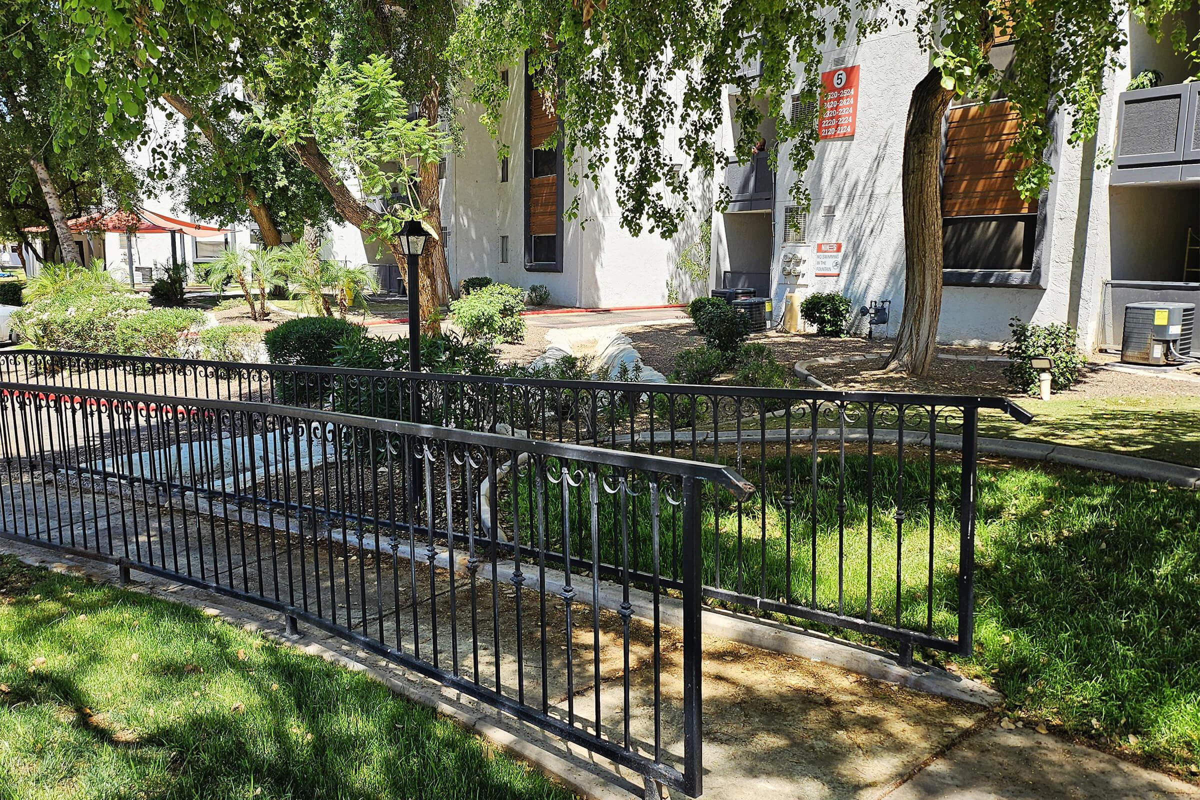 A curved black metal railing leads to a landscaped area with grass, shrubs, and trees. In the background, a building with light-colored walls and large windows is visible. The scene is bright and sunny, with a touch of greenery around the walkway and railing.