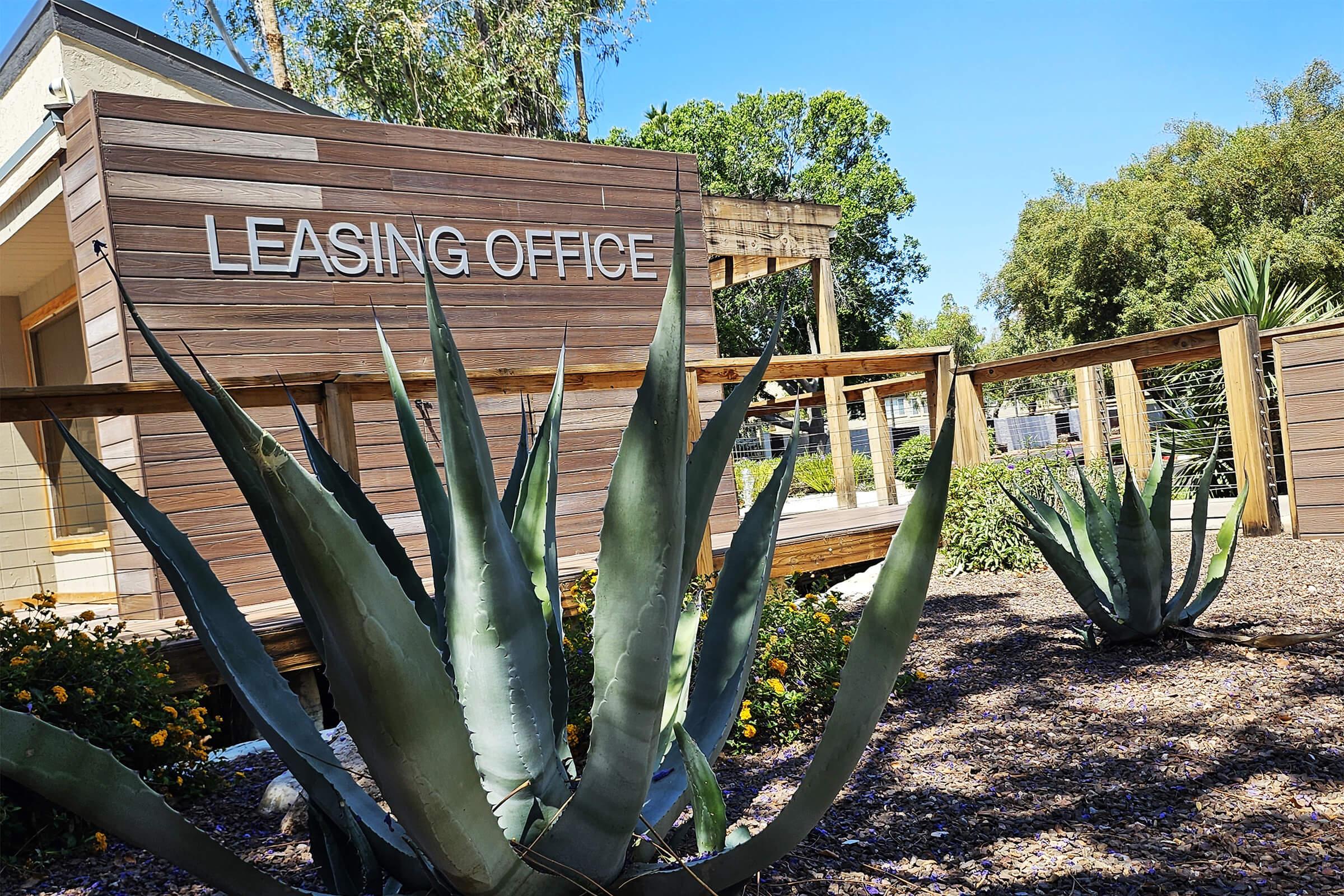 A leasing office building with a wooden facade and the words "LEASING OFFICE" prominently displayed. In the foreground, large agave plants are visible, surrounded by mulch and small flowers. The scene is set on a sunny day with clear blue skies and greenery in the background.