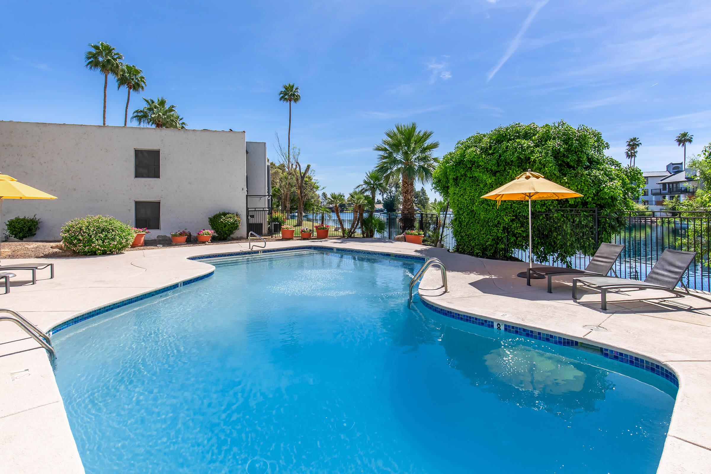 A well-maintained swimming pool surrounded by palm trees and lounge chairs. Bright yellow umbrellas provide shade, and a calm body of water is visible in the background under a clear blue sky. The setting offers a relaxing atmosphere for leisure and enjoyment.