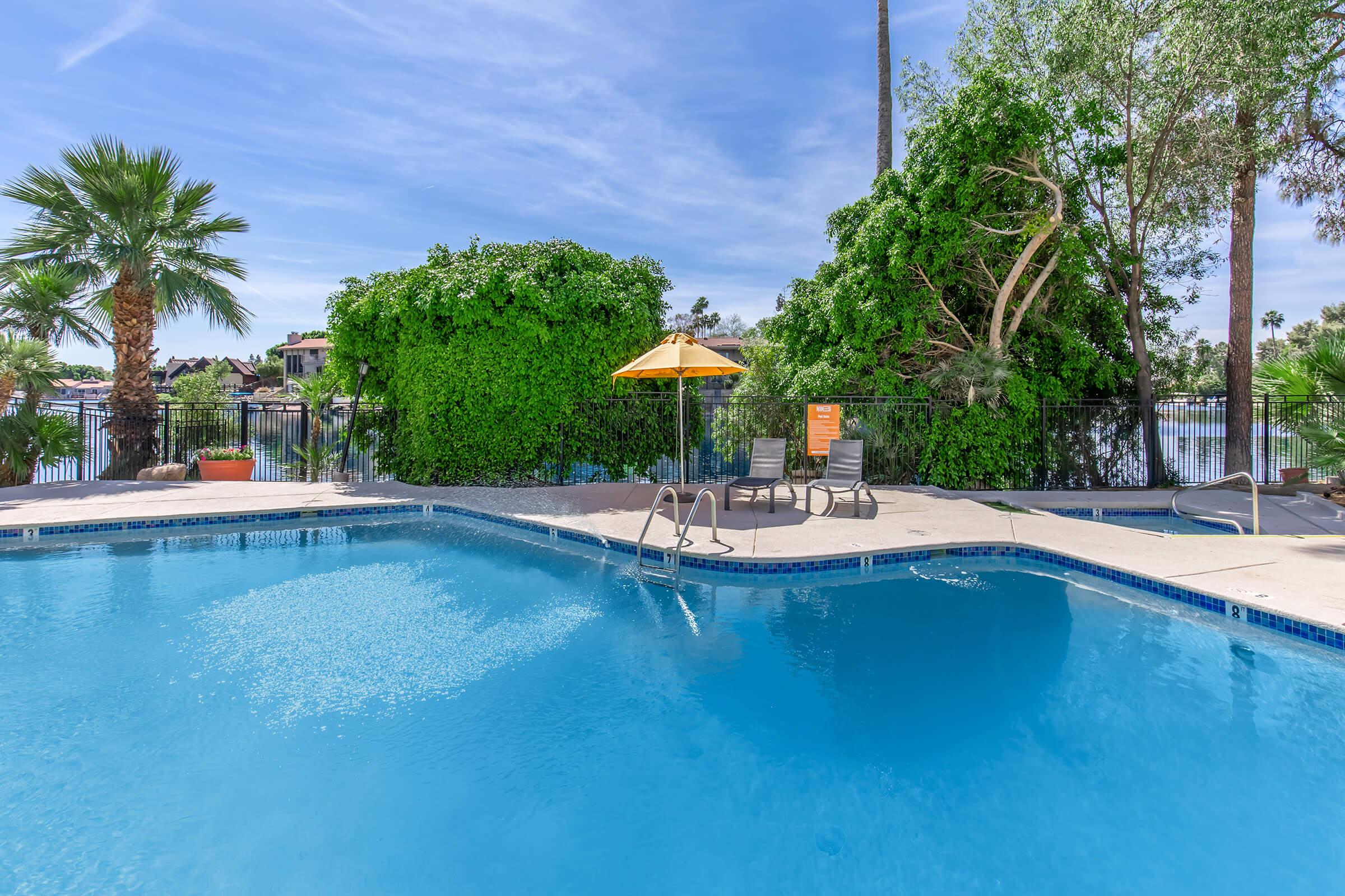 A clear blue swimming pool surrounded by lush greenery and palm trees. There is a yellow umbrella providing shade over a lounge chair. The pool area is well-maintained, with a calm lake visible in the background and a sunny sky, creating a relaxing outdoor atmosphere.