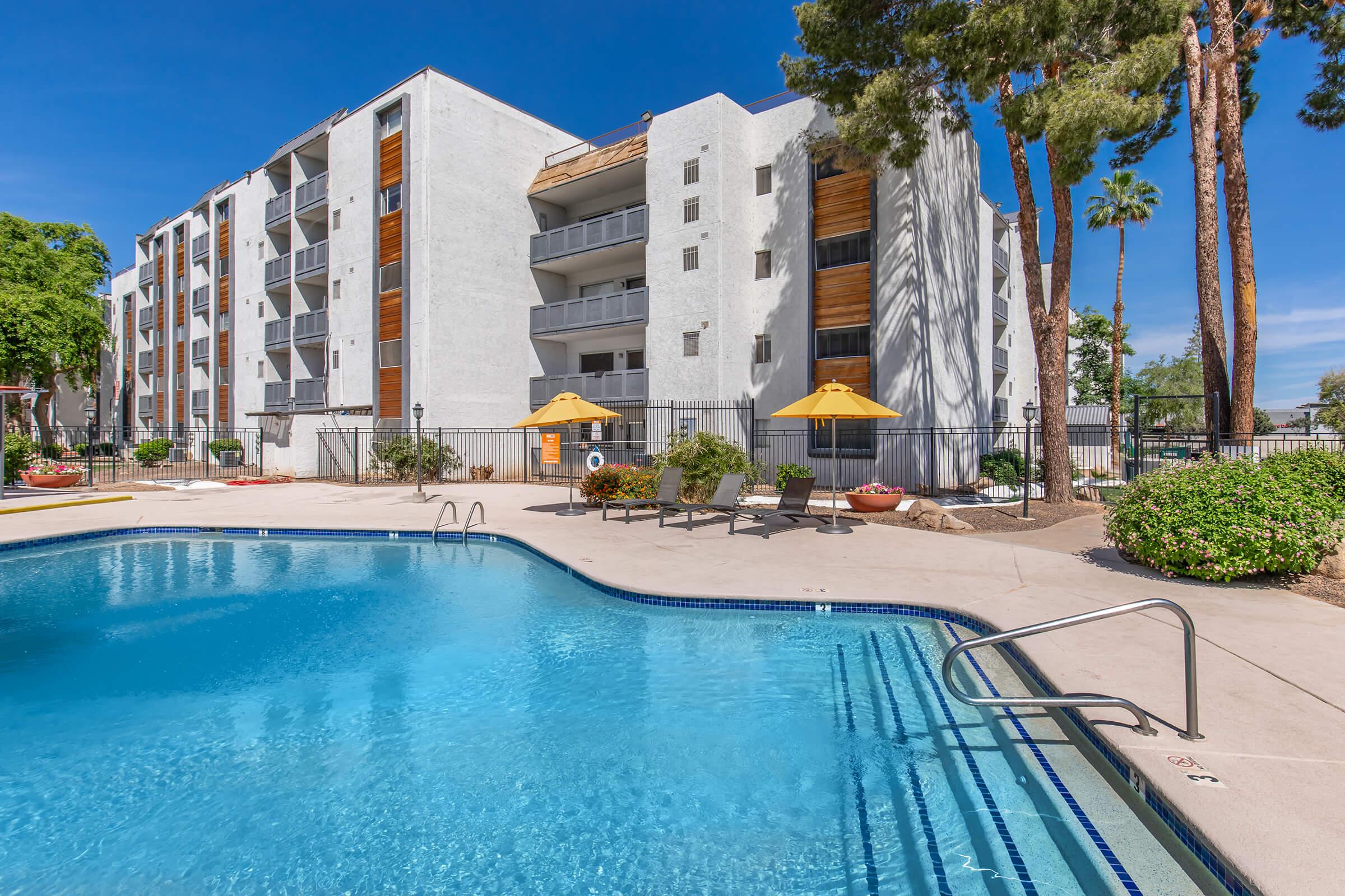 Swimming pool surrounded by a sunny courtyard featuring lounge chairs and umbrellas. In the background, a modern apartment building with balconies and palm trees. Clear blue skies enhance the inviting atmosphere of the recreational space.