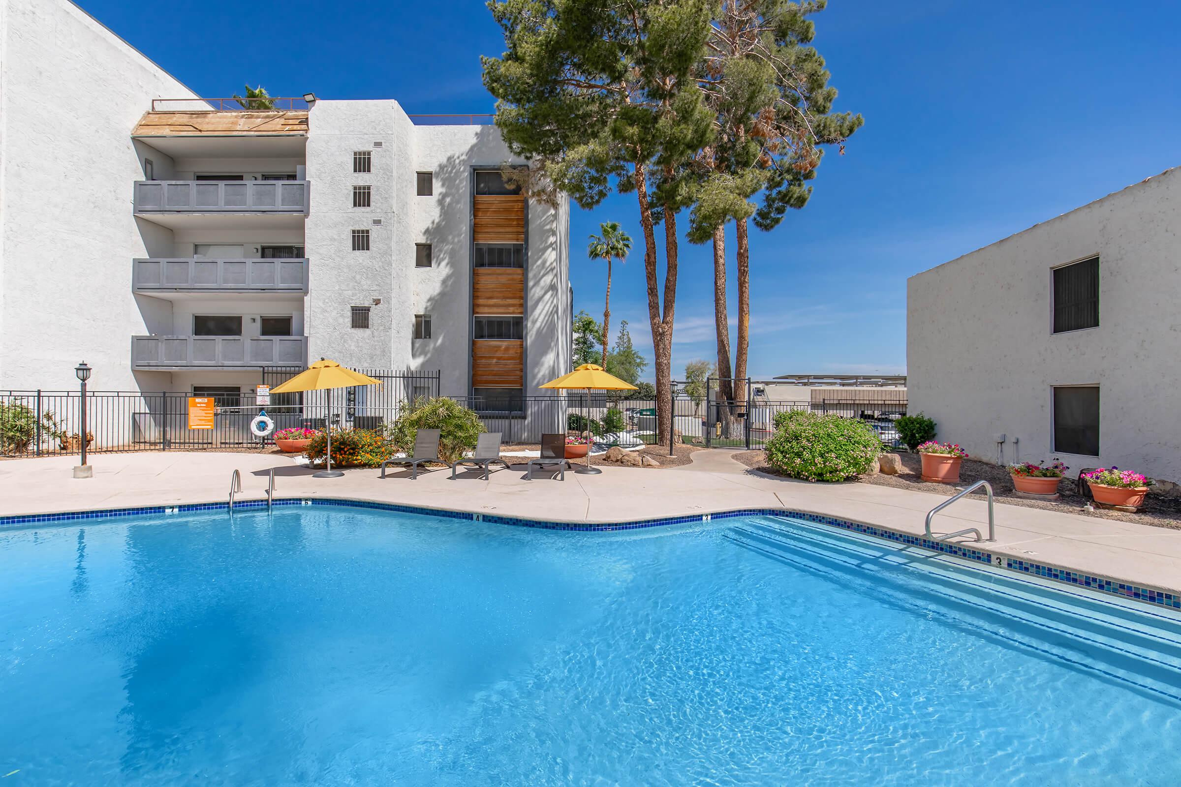 A serene outdoor pool area featuring clear blue water, surrounded by a landscaped patio with lounge chairs and yellow umbrellas. In the background, a white multi-story building with balconies and tall palm trees, under a clear blue sky, contributes to a relaxing ambiance.