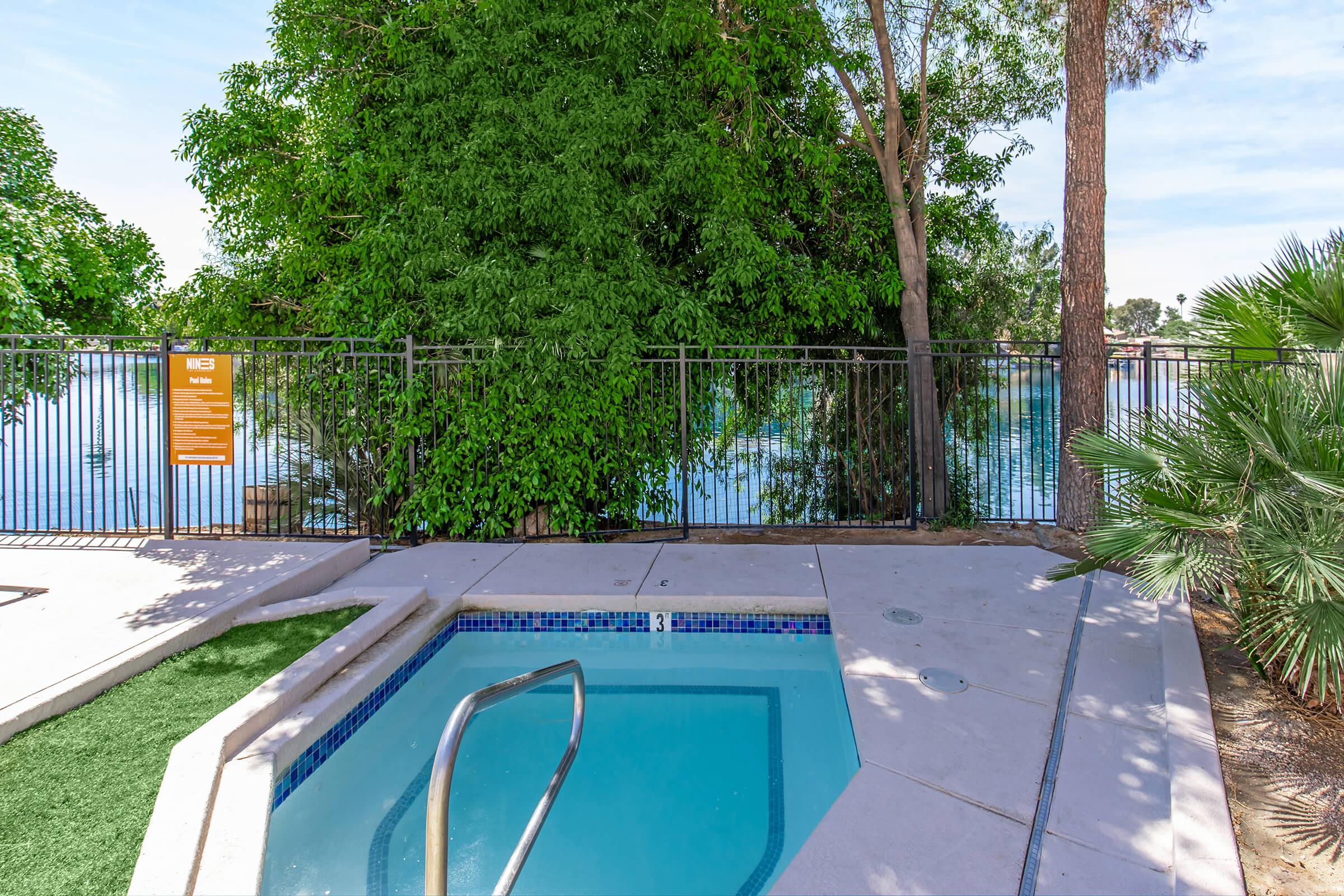 A serene pool area surrounded by lush greenery and trees, with a clear view of the water. The pool features a metal handrail for access. There is a safety fence surrounding the pool, and the ground is complemented by patches of grass. A nearby sign provides information, enhancing the tranquil atmosphere.