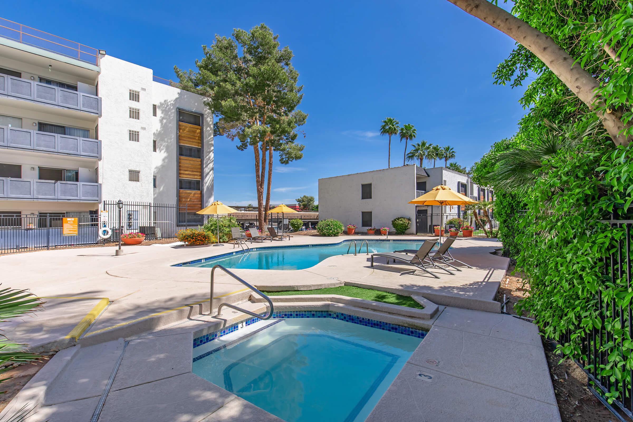 A bright outdoor swimming pool area featuring a larger pool and a smaller hot tub. Surrounding the pool are lounge chairs, umbrellas, and tropical landscaping. In the background, there is a multi-story apartment building and palm trees under a clear blue sky.