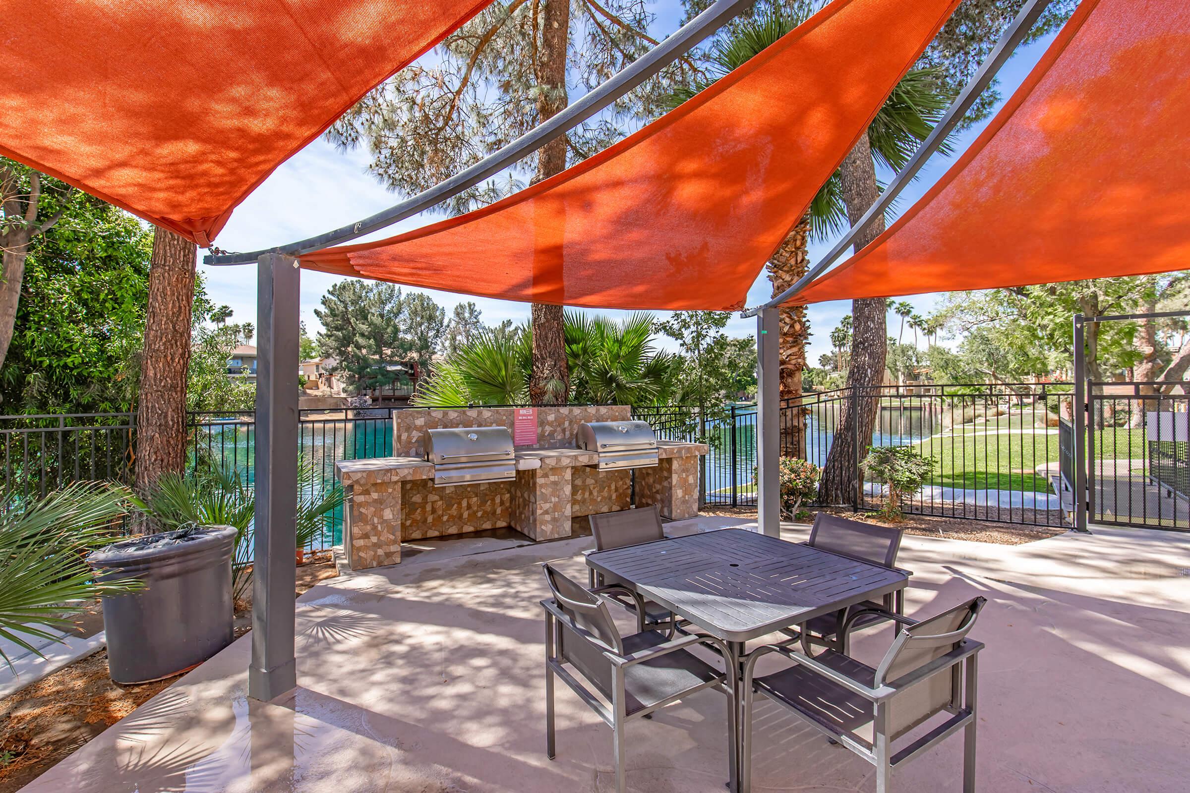 A shaded outdoor patio area with orange sail-like canopies, featuring a table with black chairs, and a grill station with two barbecue grills. Lush greenery surrounds the space, and a swimming pool is visible in the background. Ideal setting for outdoor cooking and gatherings.