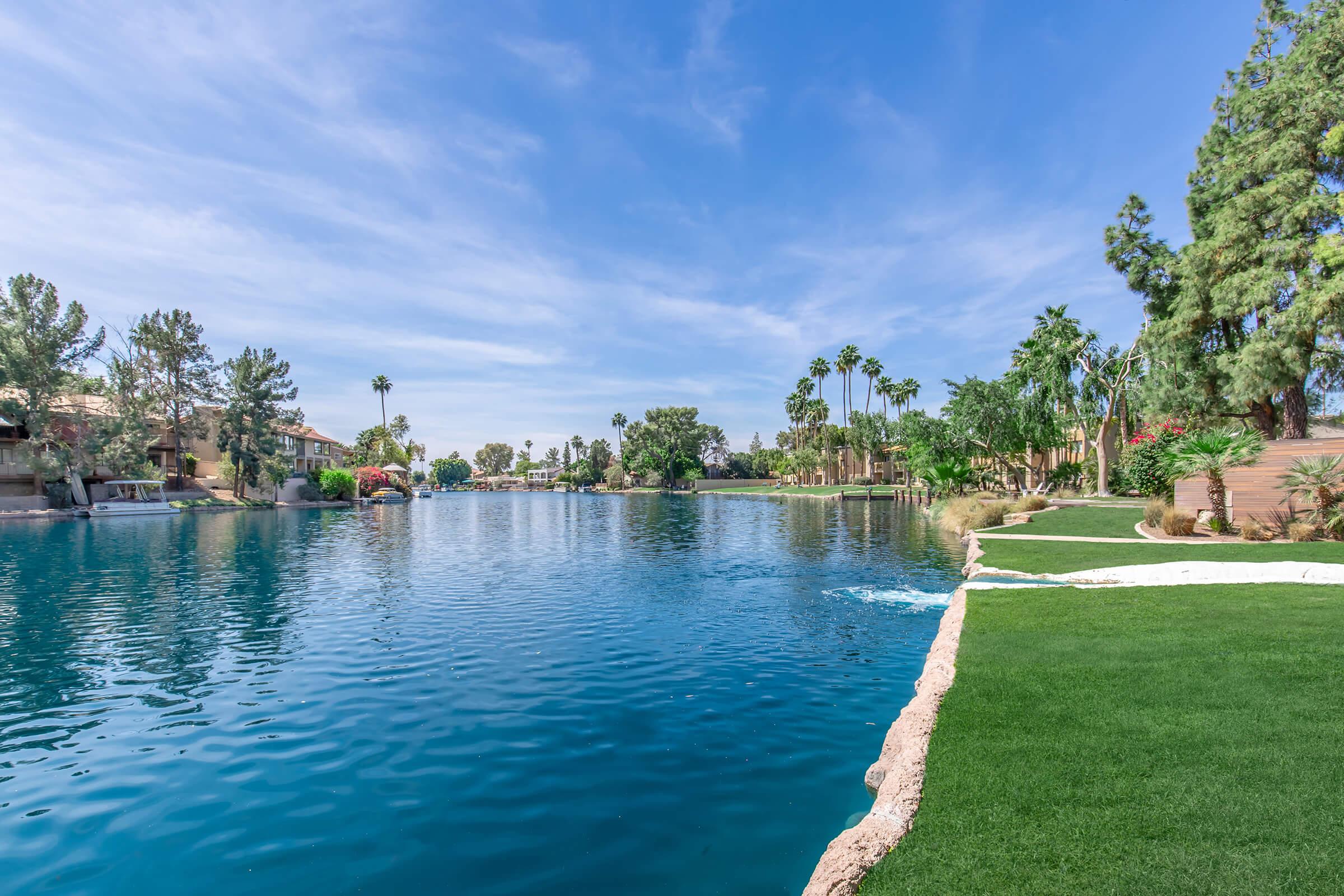 A serene lake surrounded by lush greenery and palm trees under a clear blue sky. The water reflects the landscape, creating a peaceful atmosphere. A manicured lawn edges the water, inviting relaxation and enjoyment of the natural surroundings.