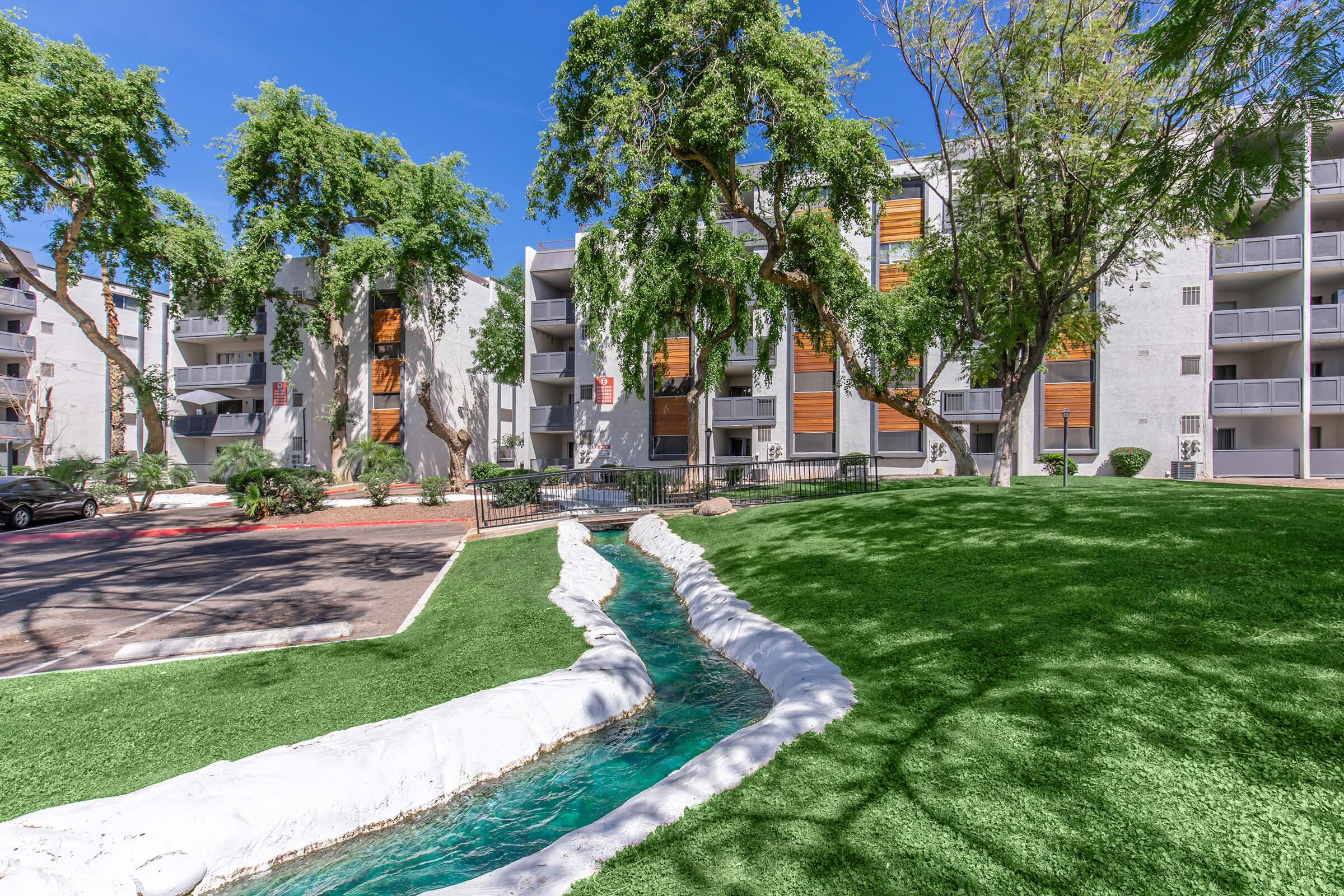 A landscaped area featuring a small, flowing stream surrounded by green grass and trees. In the background, there are modern residential buildings with wooden accents and balconies. The sky is clear and blue, creating a bright and inviting atmosphere.