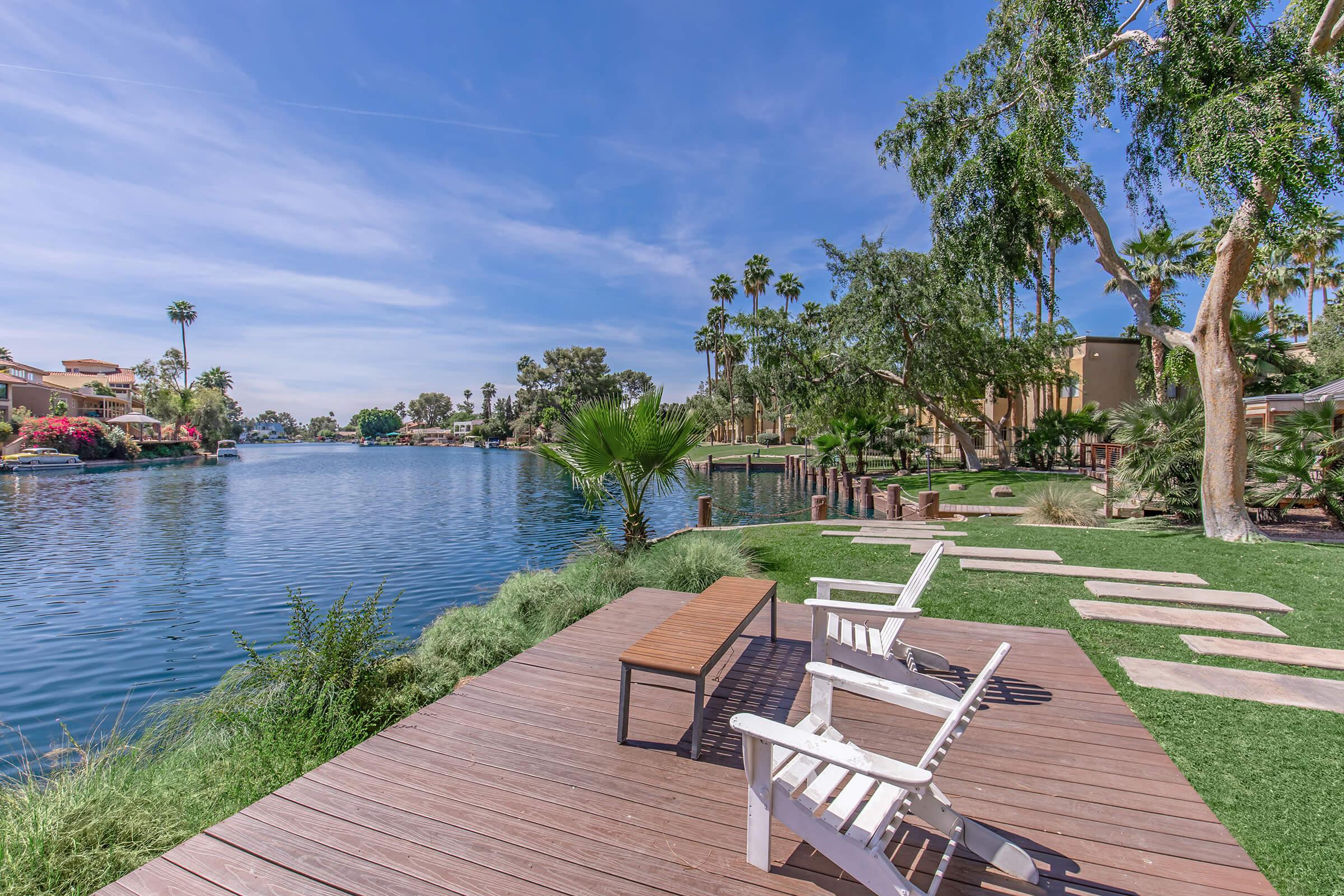 A peaceful lakeside scene featuring wooden lounge chairs on a deck, surrounded by lush greenery and palm trees. The calm water reflects the blue sky, with walking paths leading along the shore, creating a relaxing outdoor atmosphere. Ideal for leisure and enjoying nature.