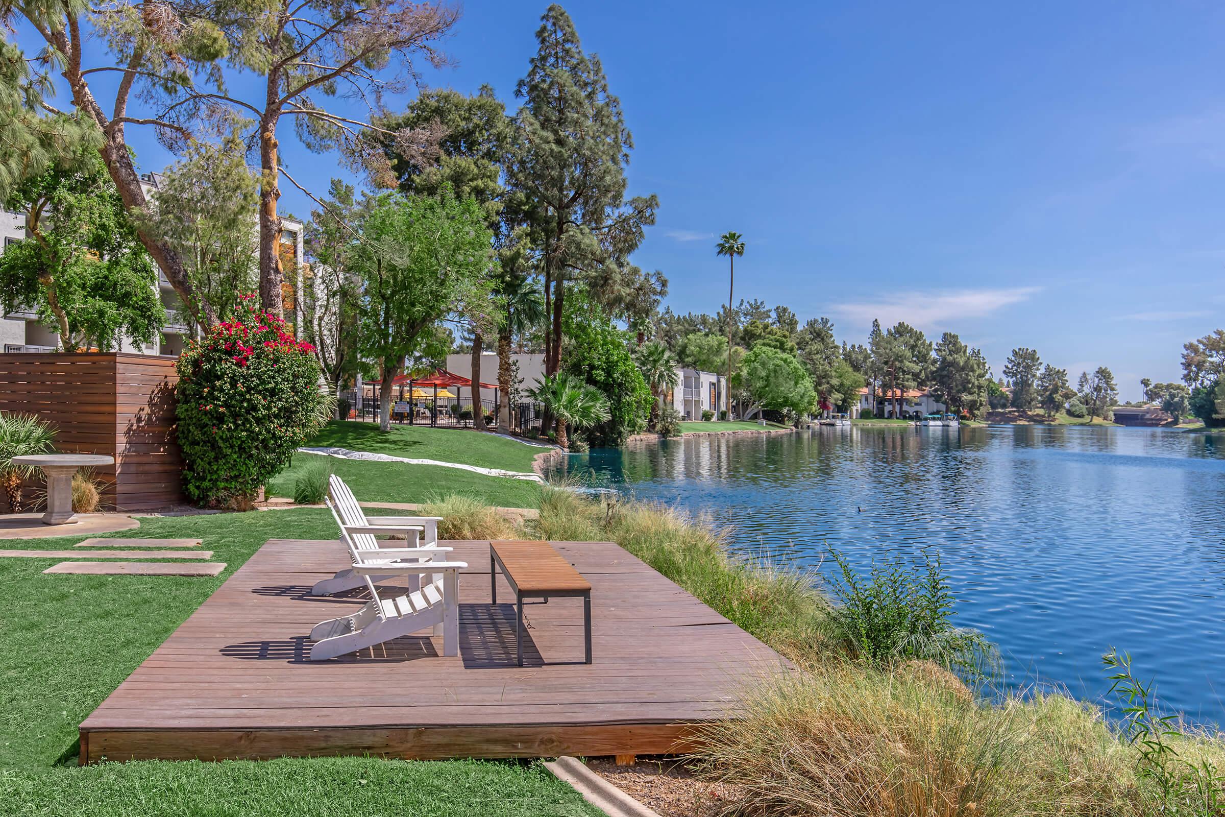 A serene lakeside view featuring a wooden deck with two white adirondack chairs and a coffee table. Lush green grass surrounds the deck, and tall trees provide shade. The calm blue water reflects the clear sky, with distant houses visible along the shore. Ideal for relaxation and enjoying nature.