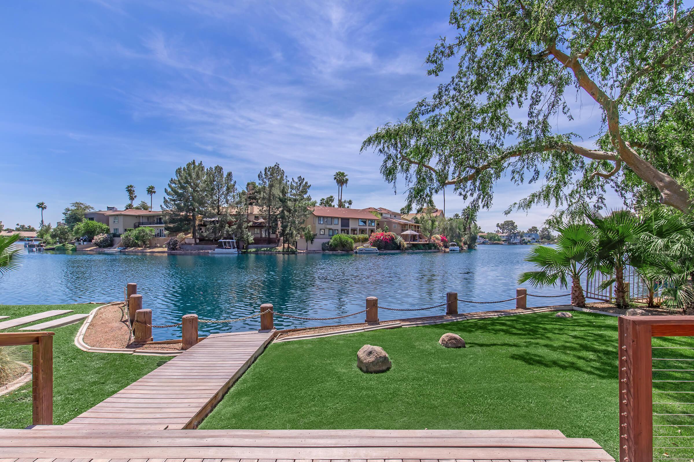 A serene lakeside view featuring a wooden walkway leading to the water, bordered by lush green grass and palm trees. In the background, residential buildings are surrounded by trees and colorful flowers, reflecting off the calm lake under a clear blue sky.