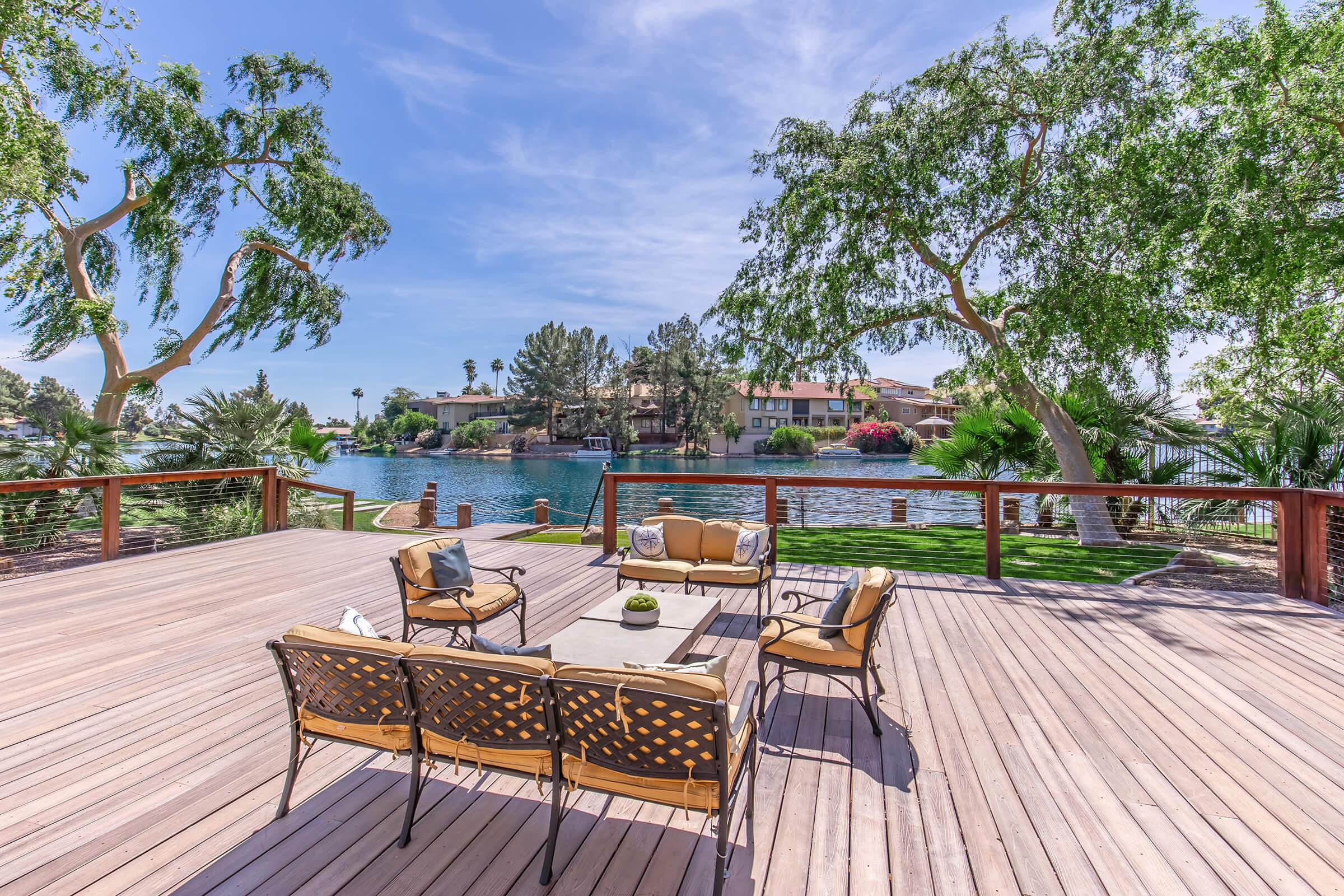A serene lakeside patio featuring comfortable outdoor seating with a coffee table, surrounded by lush greenery and palm trees. In the background, a tranquil lake reflects the blue sky and nearby houses, creating a peaceful outdoor retreat.