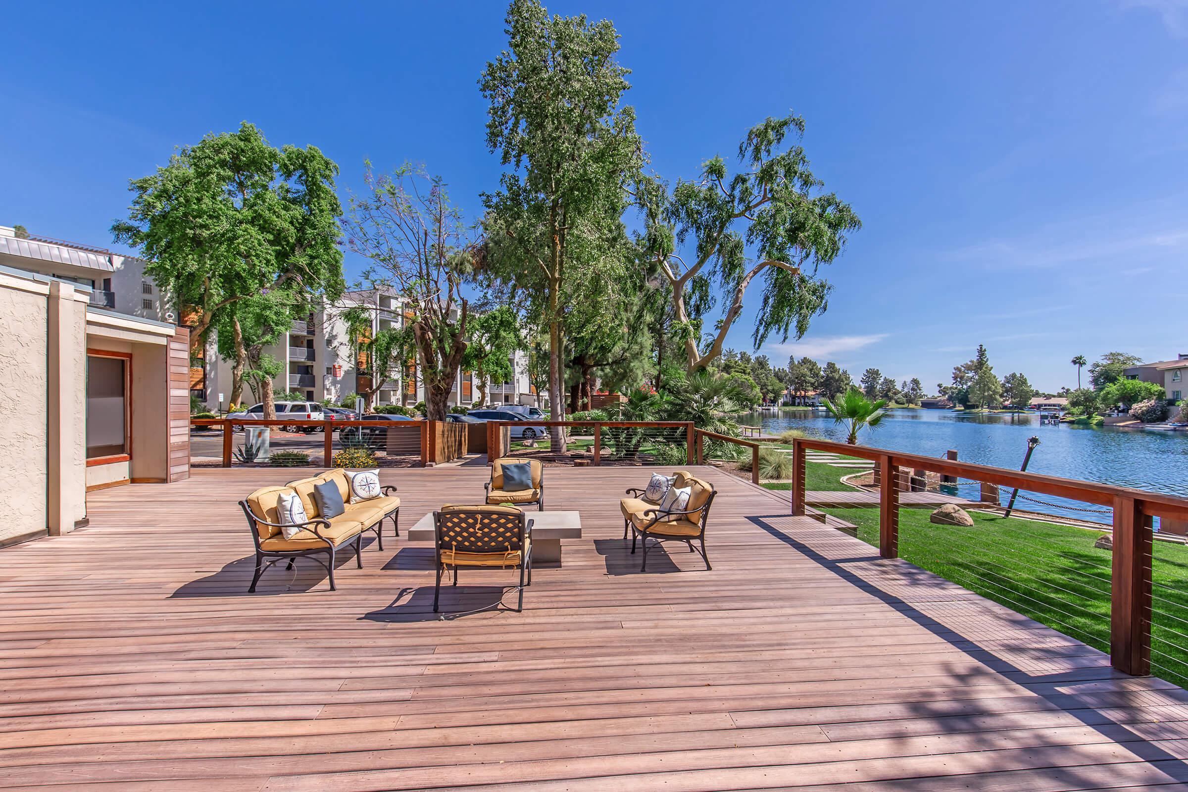 A wooden deck overlooking a serene lake, surrounded by lush greenery and trees. There are comfortable outdoor chairs and a table set up for relaxation. In the background, residential buildings can be seen along the water's edge, with blue skies and clear weather creating a tranquil atmosphere.