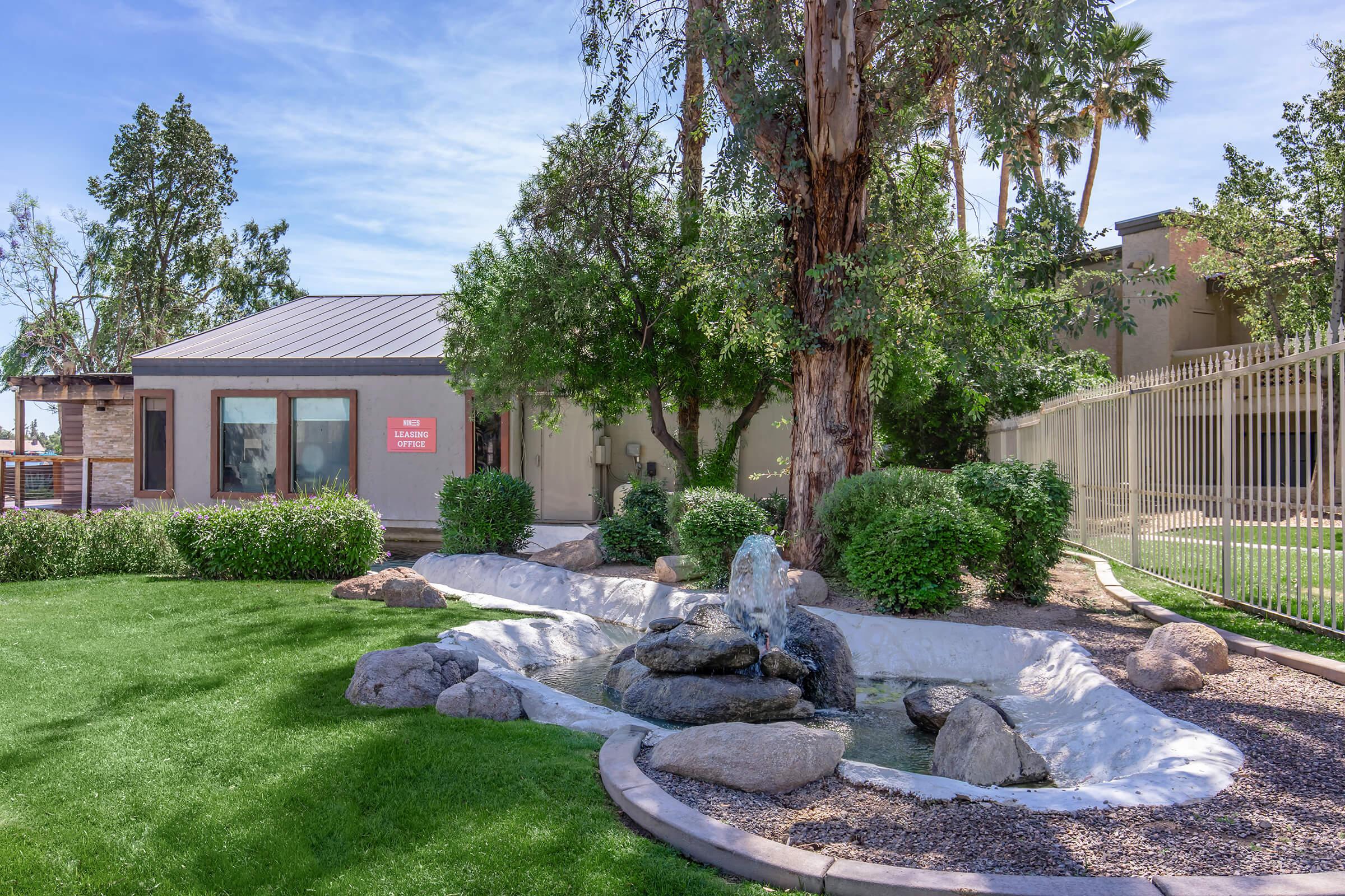 A small, landscaped area featuring a decorative outdoor fountain surrounded by rocks and greenery. In the background, there is a modern building with a sign visible, surrounded by trees and a fence. The setting appears well-maintained and inviting, ideal for relaxation.