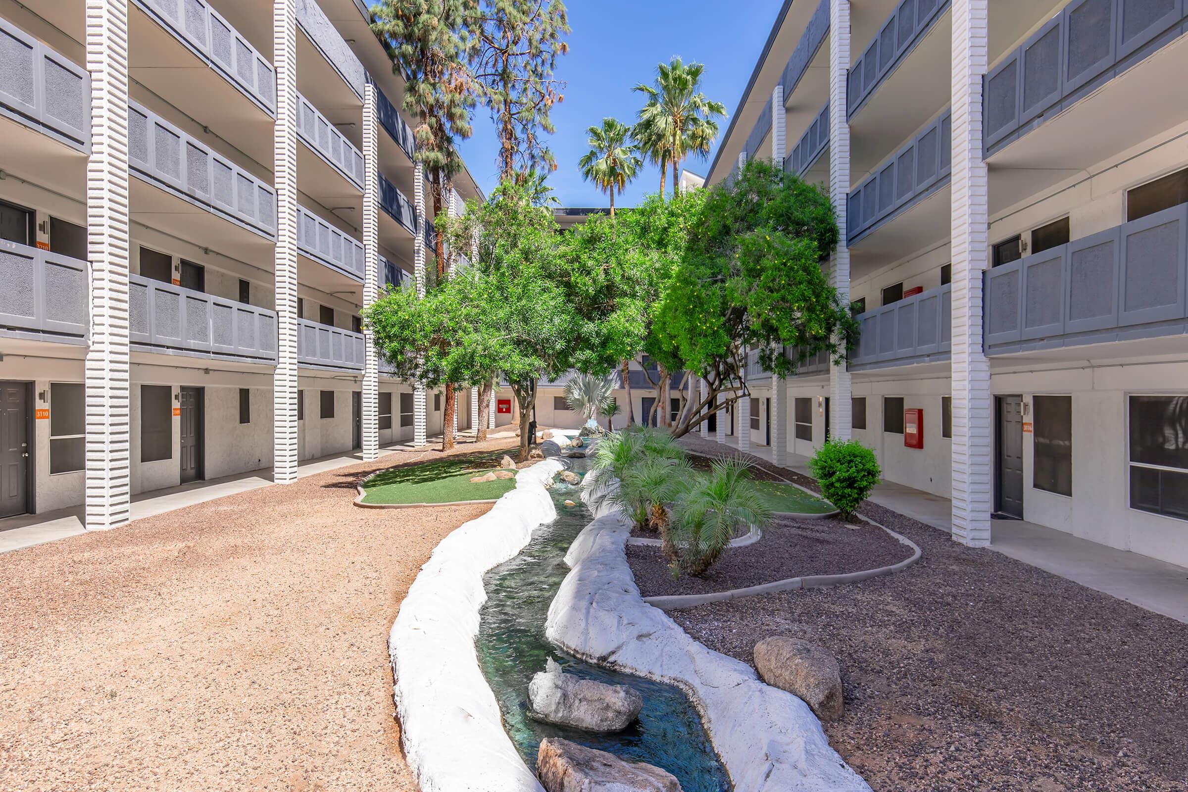 A serene courtyard featuring lush greenery and a small stream. Surrounding the area are two-story apartment buildings with balconies. The landscape includes palm trees and landscaped rocks, creating a peaceful and inviting ambiance. Bright blue sky completes the scene.