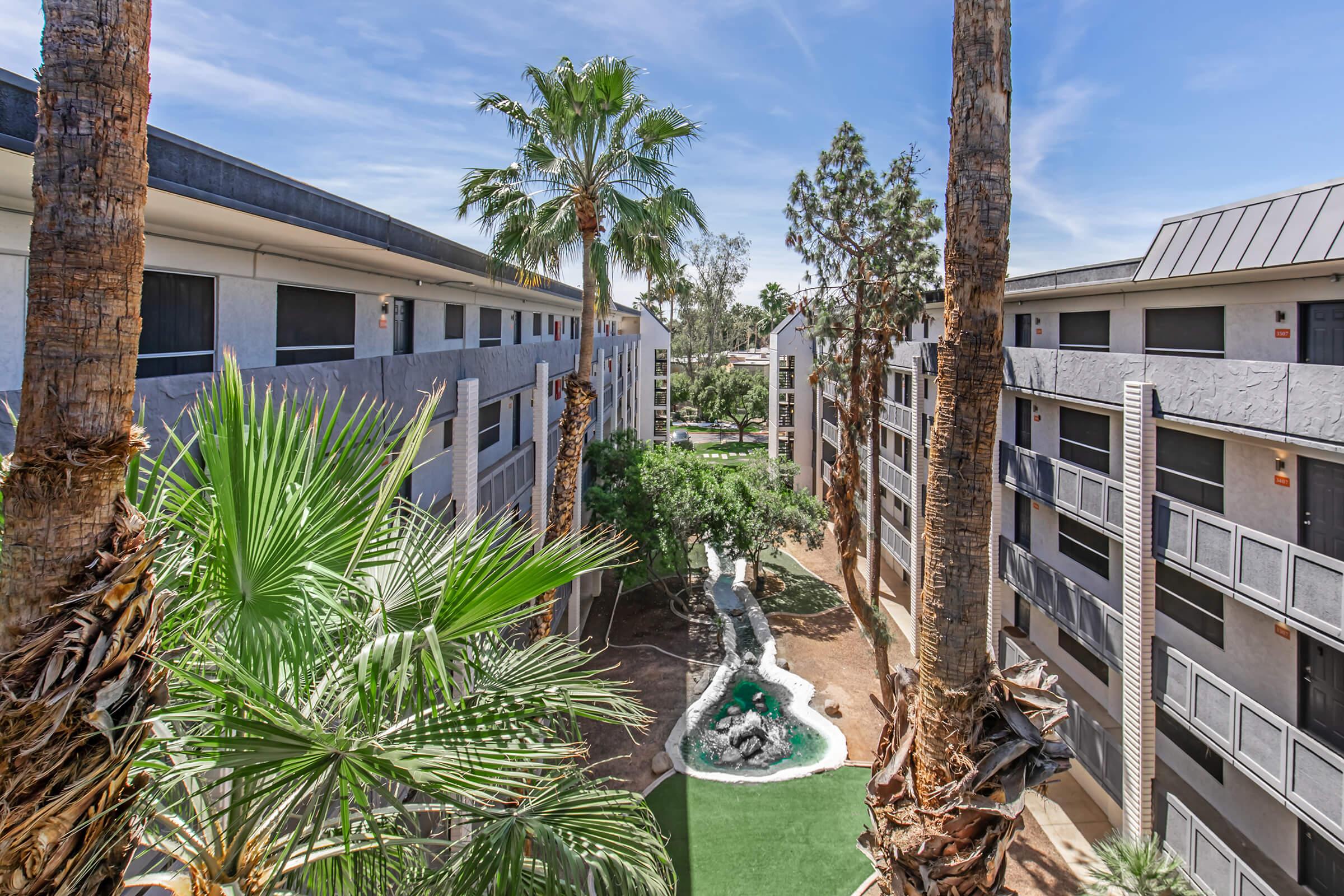 View of a modern apartment complex featuring palm trees, green landscaping, and a small water feature. The buildings are multi-storied with balconies, surrounded by lush greenery and clear blue skies. A vibrant outdoor space enhances the community feel, inviting residents to enjoy the area.