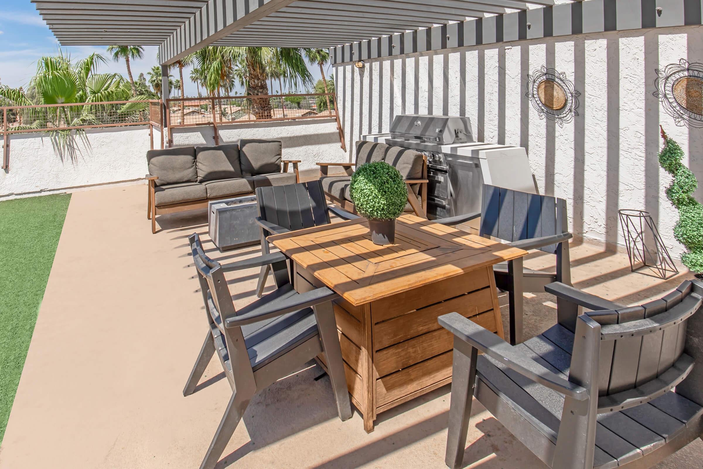 Outdoor patio area with a wooden table at the center surrounded by several chairs. A sleek gray sofa is positioned to one side, and there is a barbecue grill in the background. The space features green grass and palm trees, complemented by a pergola providing partial shade.