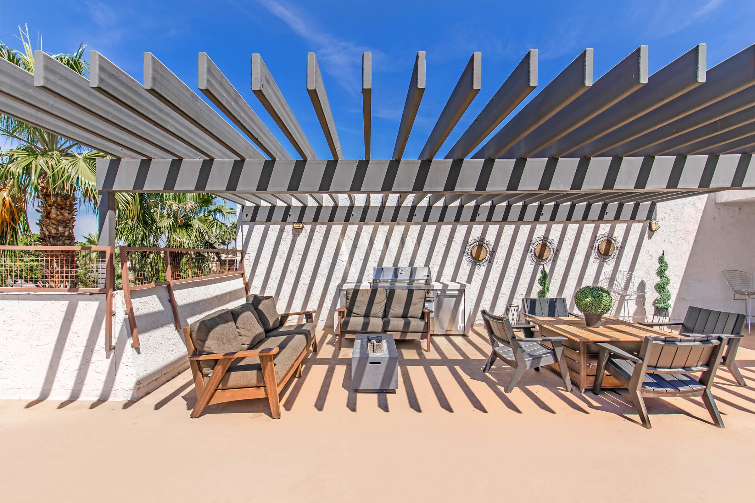A modern outdoor patio featuring a shaded area with horizontal metal beams casting shadows onto the ground. The space includes comfortable seating with cushions, a wooden dining table, decorative plants, and a grilling area surrounded by palm trees, all set against a clear blue sky.