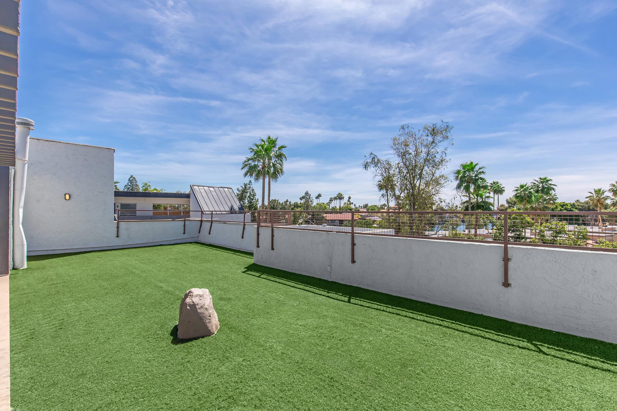 A spacious outdoor terrace featuring artificial grass, surrounded by palm trees under a bright blue sky. There is a large rock placed on the lawn and a railing along one side of the terrace, providing an open view of the surroundings.