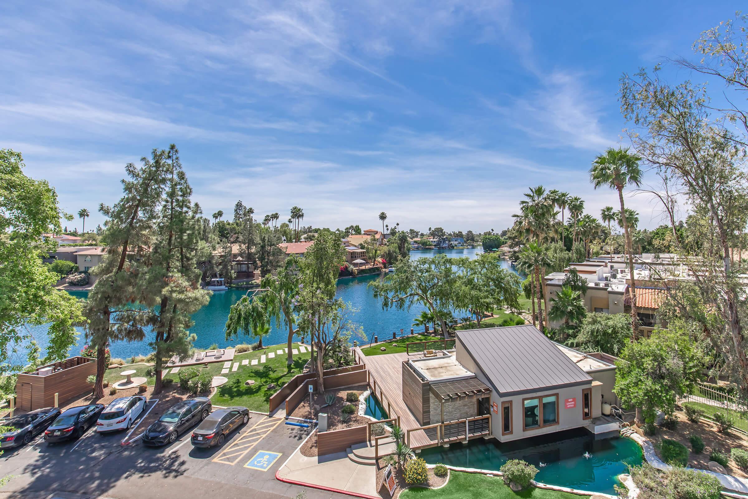 A scenic view of a waterfront area featuring lush trees, palm trees, and houses along the shoreline. The water reflects the blue sky with wispy clouds, while a modern building is visible in the foreground, surrounded by well-maintained landscaping and parked vehicles.