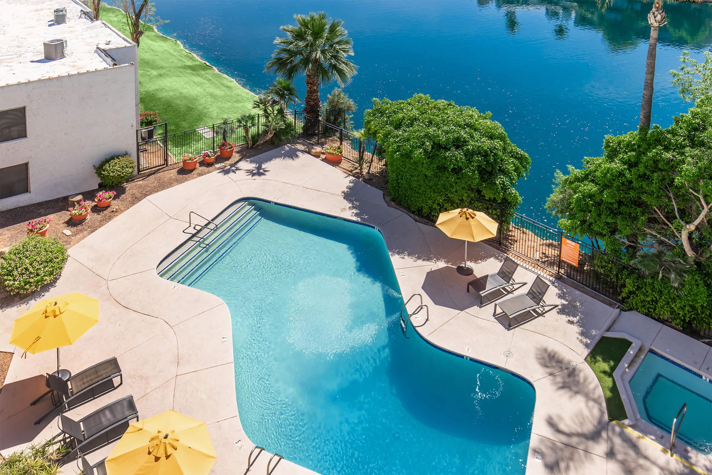 Aerial view of a tranquil swimming pool area surrounded by lush greenery and palm trees. The pool features a smooth, curved shape with lounge chairs and yellow umbrellas. Nearby, a shimmering body of water reflects the surroundings, creating a serene atmosphere.