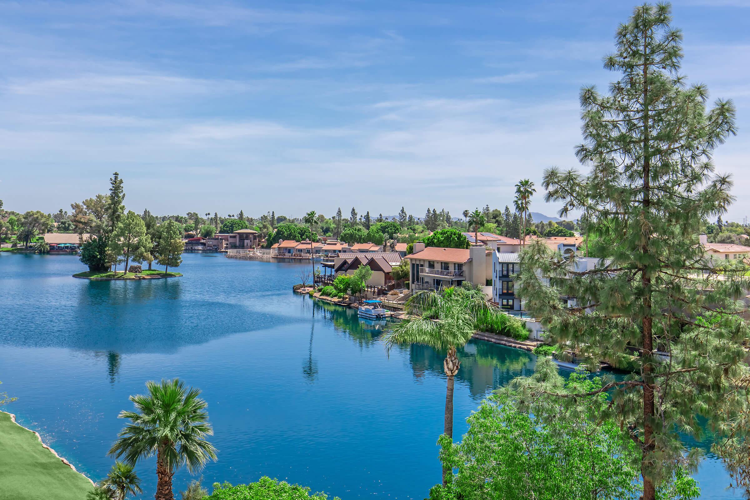 A scenic view of a calm lake surrounded by lush greenery and palm trees, with residential buildings and villas along the shoreline. The clear blue sky reflects on the water, creating a tranquil atmosphere. Small islands with trees are visible in the lake, enhancing the picturesque landscape.