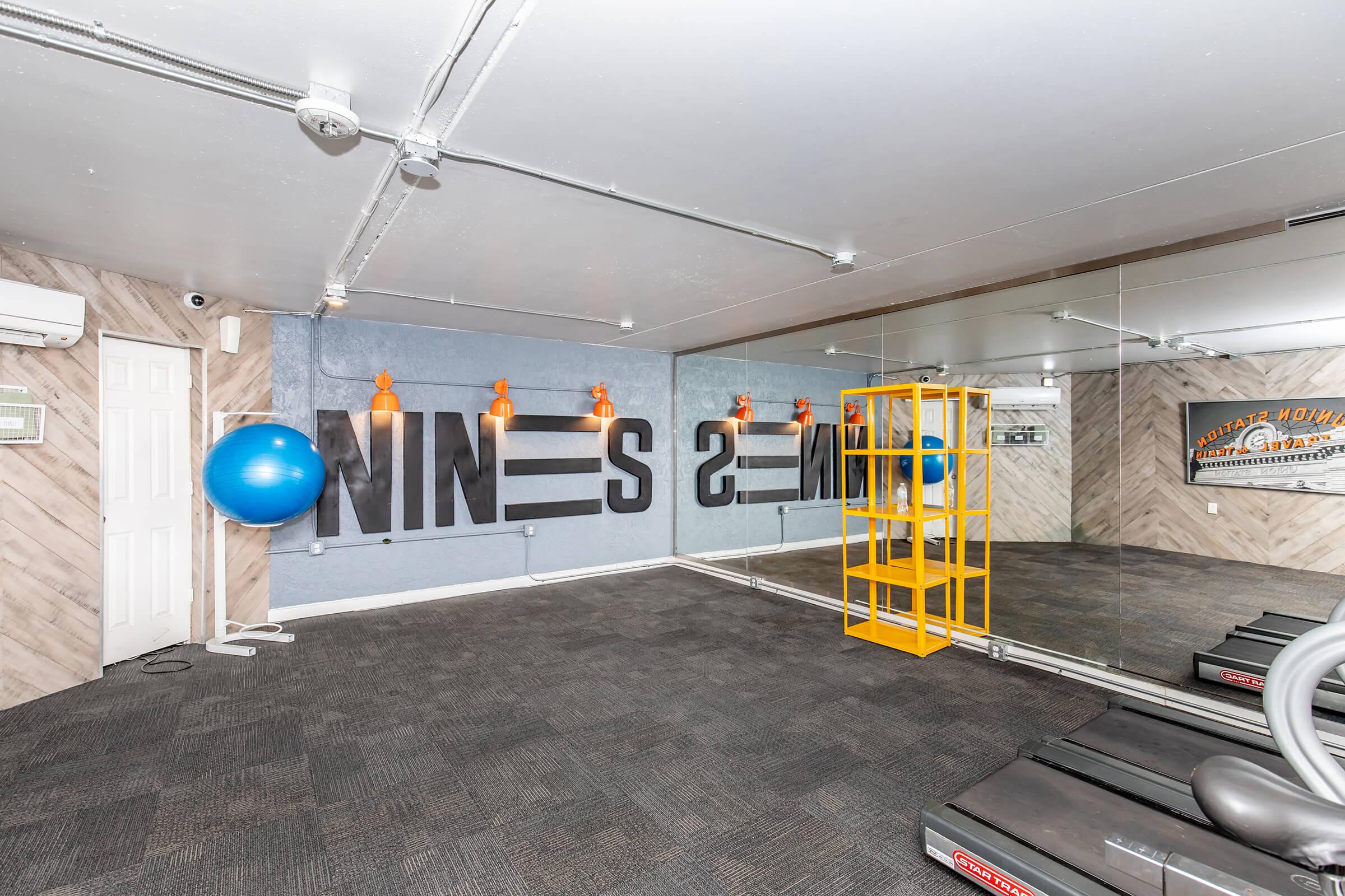 A spacious workout room featuring a large mirror, a blue exercise ball, and a yellow shelving unit. The walls are decorated with motivational phrases and a fitness emblem. The flooring is carpeted, and there are also air conditioning vents visible on the walls.