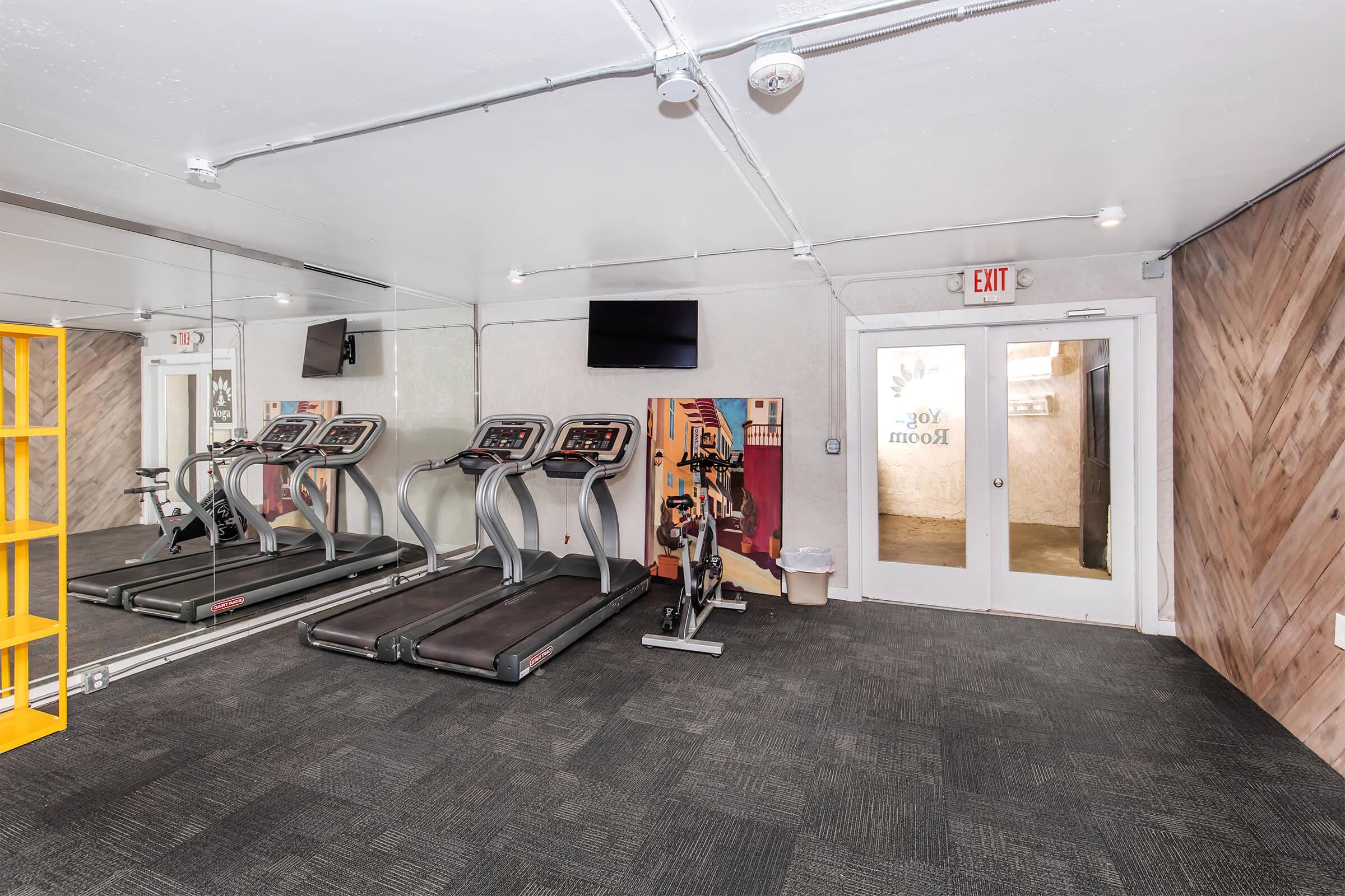 A modern gym area featuring two treadmills, a large mirror, and two flat-screen TVs mounted on the wall. The space includes a wooden accent wall and a doorway labeled "Yoga Room." The flooring is dark carpet, and the overall ambiance is bright and inviting.
