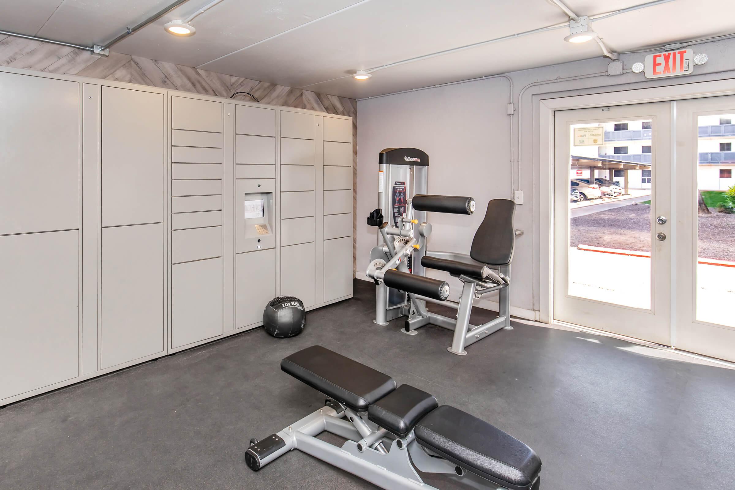 Interior view of a fitness room featuring exercise equipment, including a weight machine and a bench, alongside storage lockers. Large windows provide natural light, and an exit door leads outside. The space has a modern design with gray flooring and walls.