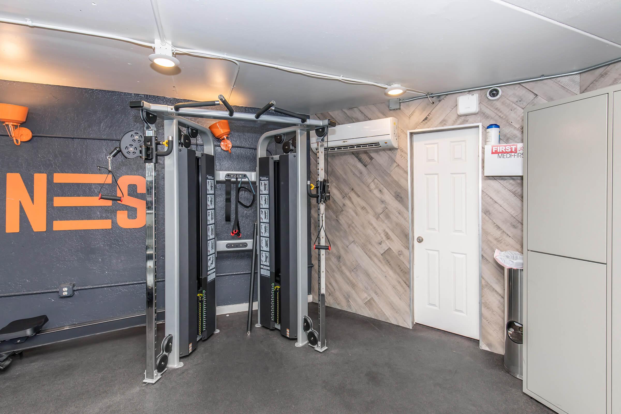 A modern gym space featuring dual cable machines, a wall with the word "Fitness," and gray wood paneling. The room is well-lit with overhead lights and includes a door, an air conditioning unit, and a trash bin in the corner.