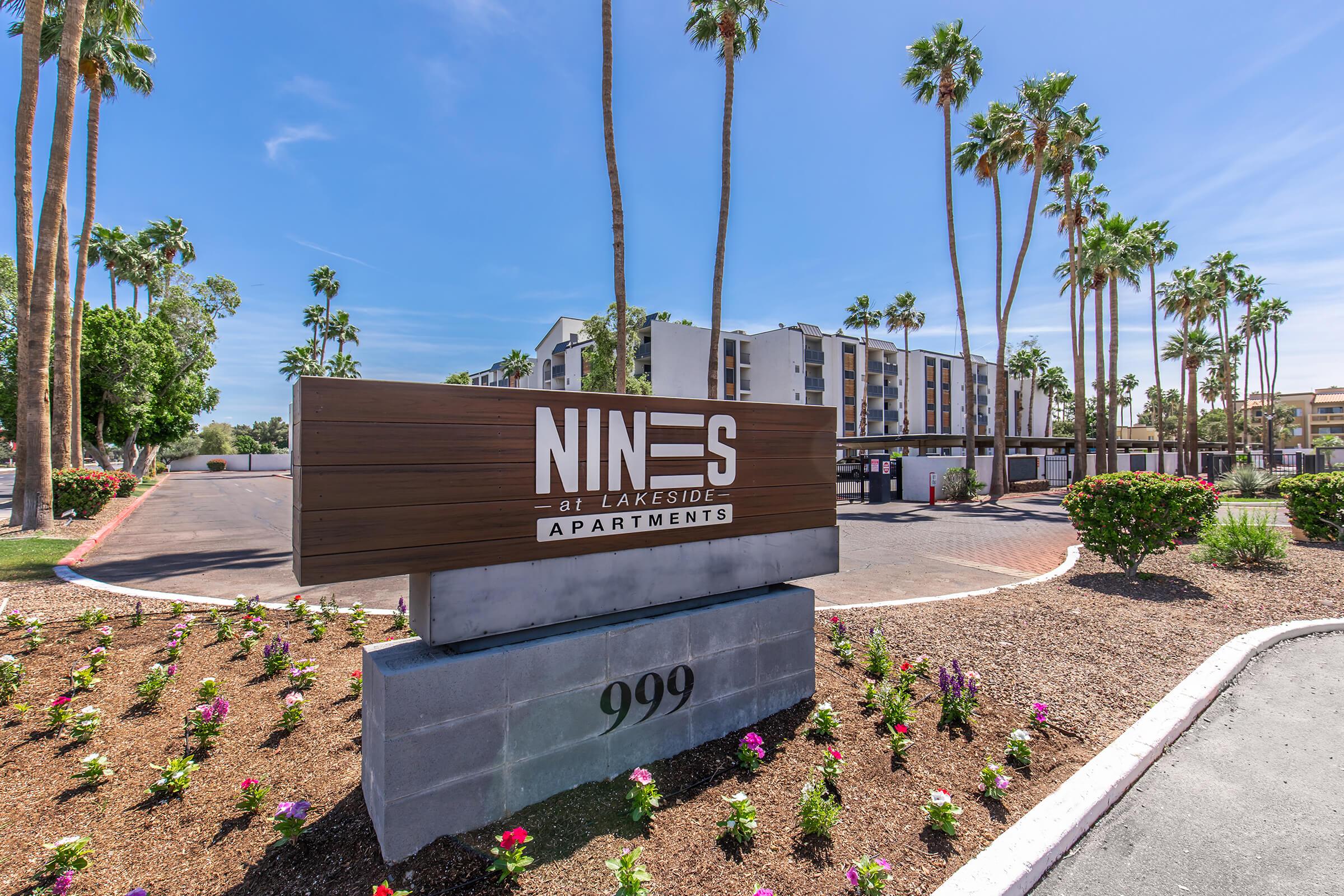 Sign for "NINES at Lakeside Apartments" with landscaping and palm trees in the foreground, set against a blue sky. The building in the background has a modern architectural design. Address: 999.