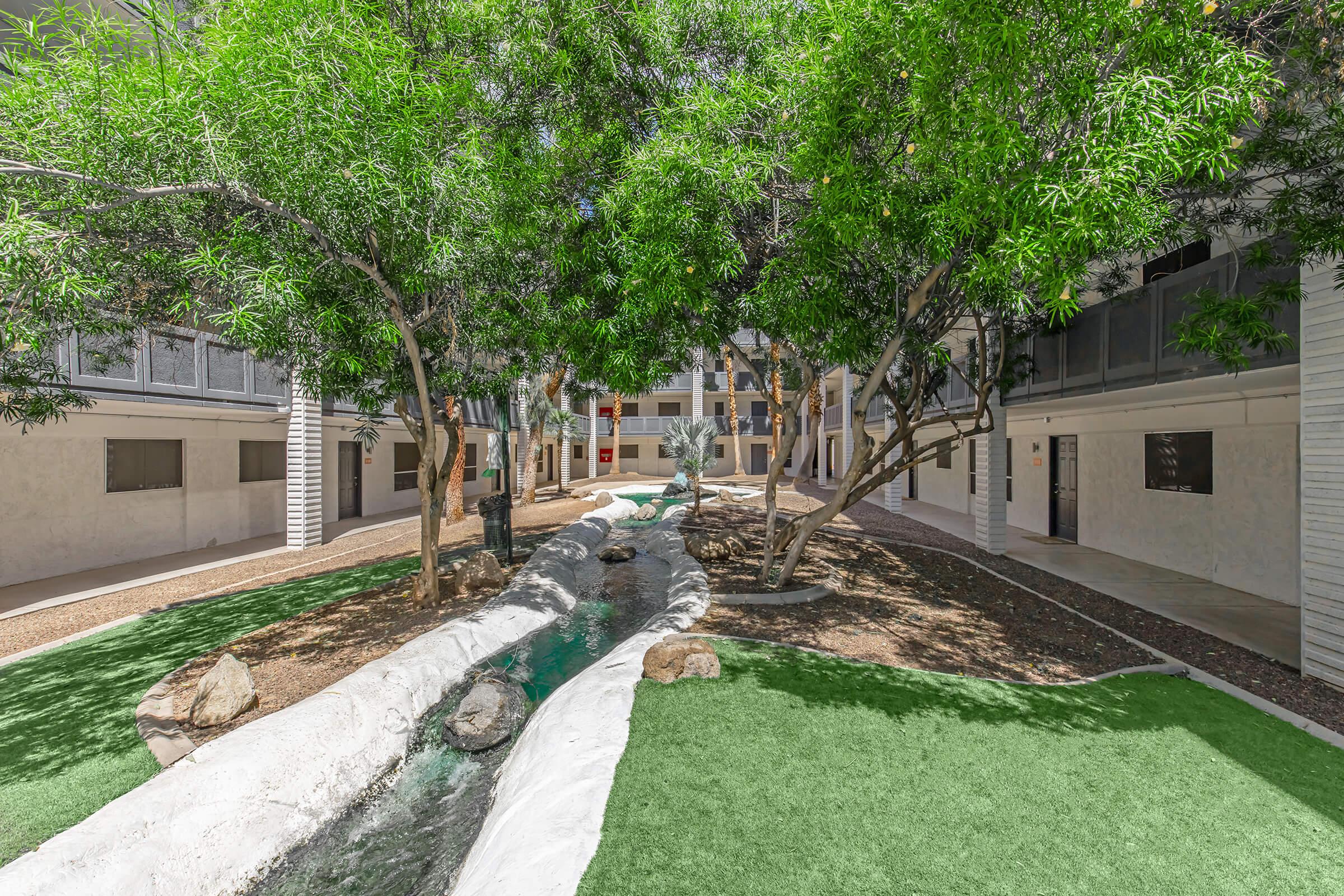 A serene courtyard featuring lush green grass, trees, and a small flowing stream. The area is surrounded by white buildings with balconies, creating a peaceful outdoor space for relaxation. Sunlight filters through the leaves, enhancing the tranquil atmosphere.